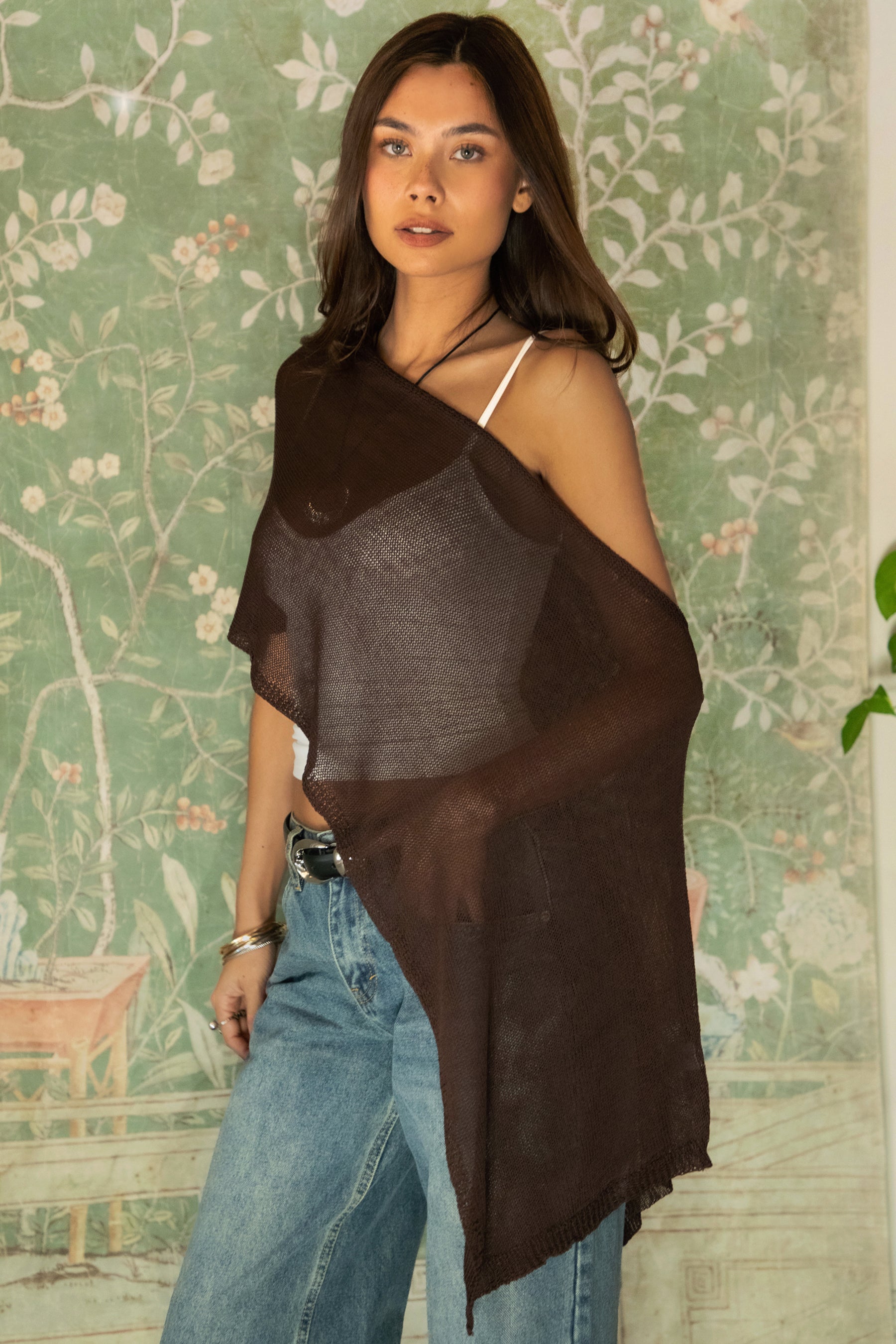 Woman wearing TIC TOC TC31873 SOLEI lightweight open sheer sweater knit poncho in BROWN, featuring a wide off-shoulder neckline, airy open knit texture, relaxed draped poncho silhouette, and asymmetrical hem, styled over a white cami with light wash denim for a western-casual summer layering look.