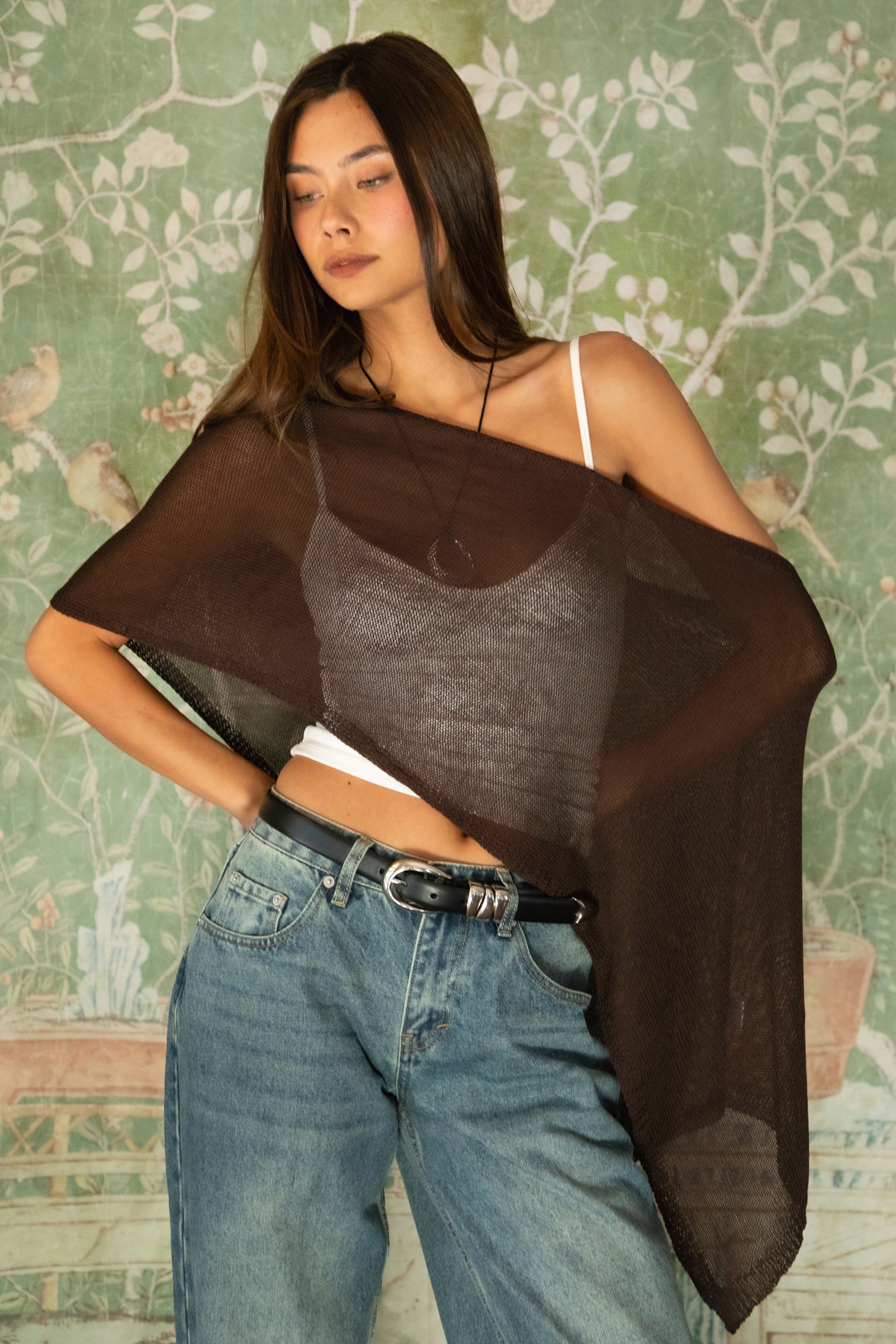 Woman wearing TIC TOC TC31873 SOLEI lightweight open sheer sweater knit poncho in BROWN, featuring a wide off-shoulder neckline, airy open knit texture, relaxed draped poncho silhouette, and asymmetrical hem, styled over a white cami with light wash denim for a western-casual summer layering look.