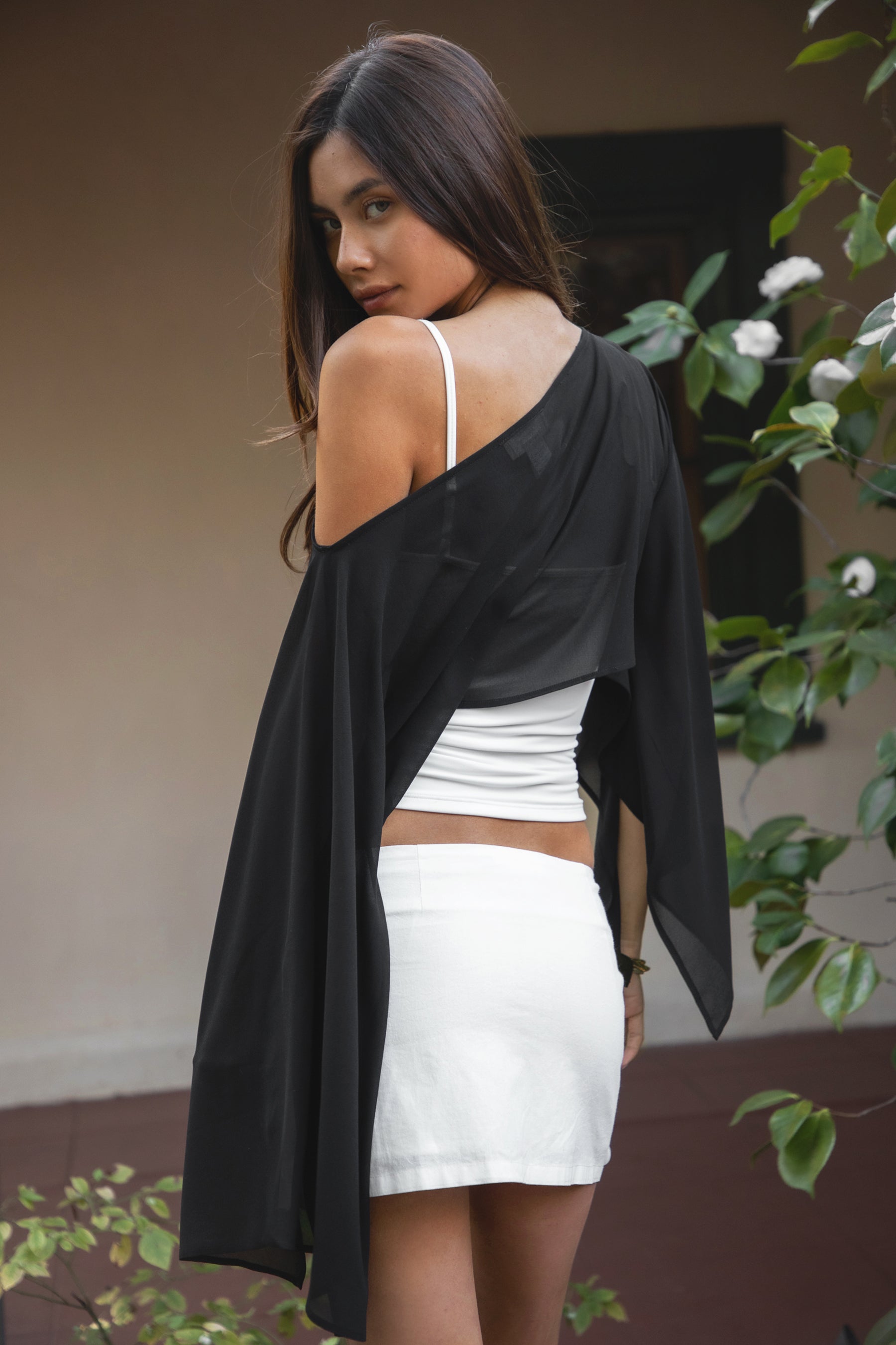 Woman wearing TIC TOC TC31933-ELYSIA ELYSIA chiffon smooth sheer flowy cape top featuring a wide boat neckline, off-shoulder cape-style silhouette with draped flare long sleeves, cropped hem, and lightweight airy chiffon fabric, styled over a fitted bralette and high-waisted bottoms for a boho chic resort-ready look.