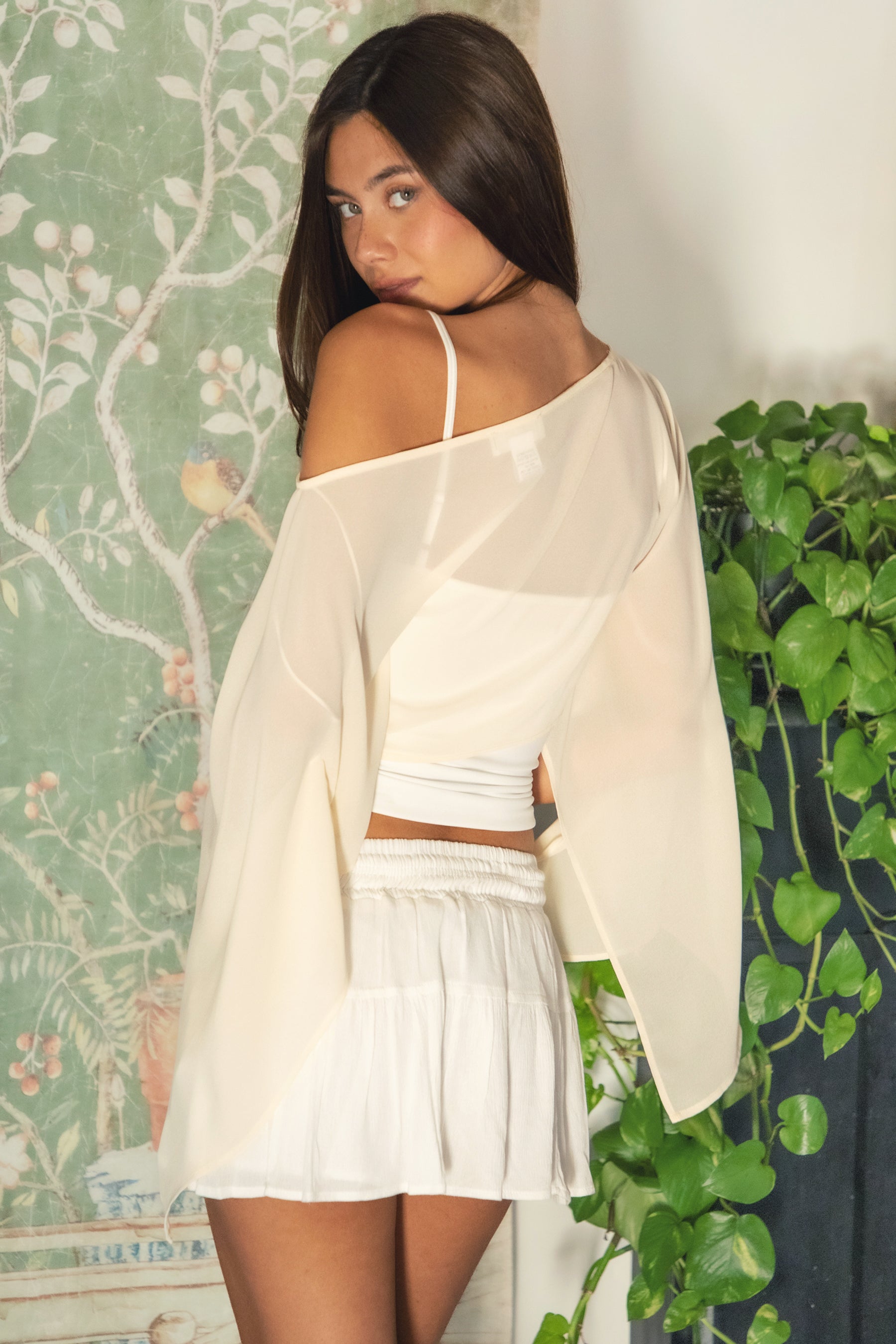 Woman wearing TIC TOC TC31933-ELYSIA ELYSIA chiffon smooth sheer flowy cape top featuring a wide boat neckline, off-shoulder cape-style silhouette with draped flare long sleeves, cropped hem, and lightweight airy chiffon fabric, styled over a fitted bralette and high-waisted bottoms for a boho chic resort-ready look.