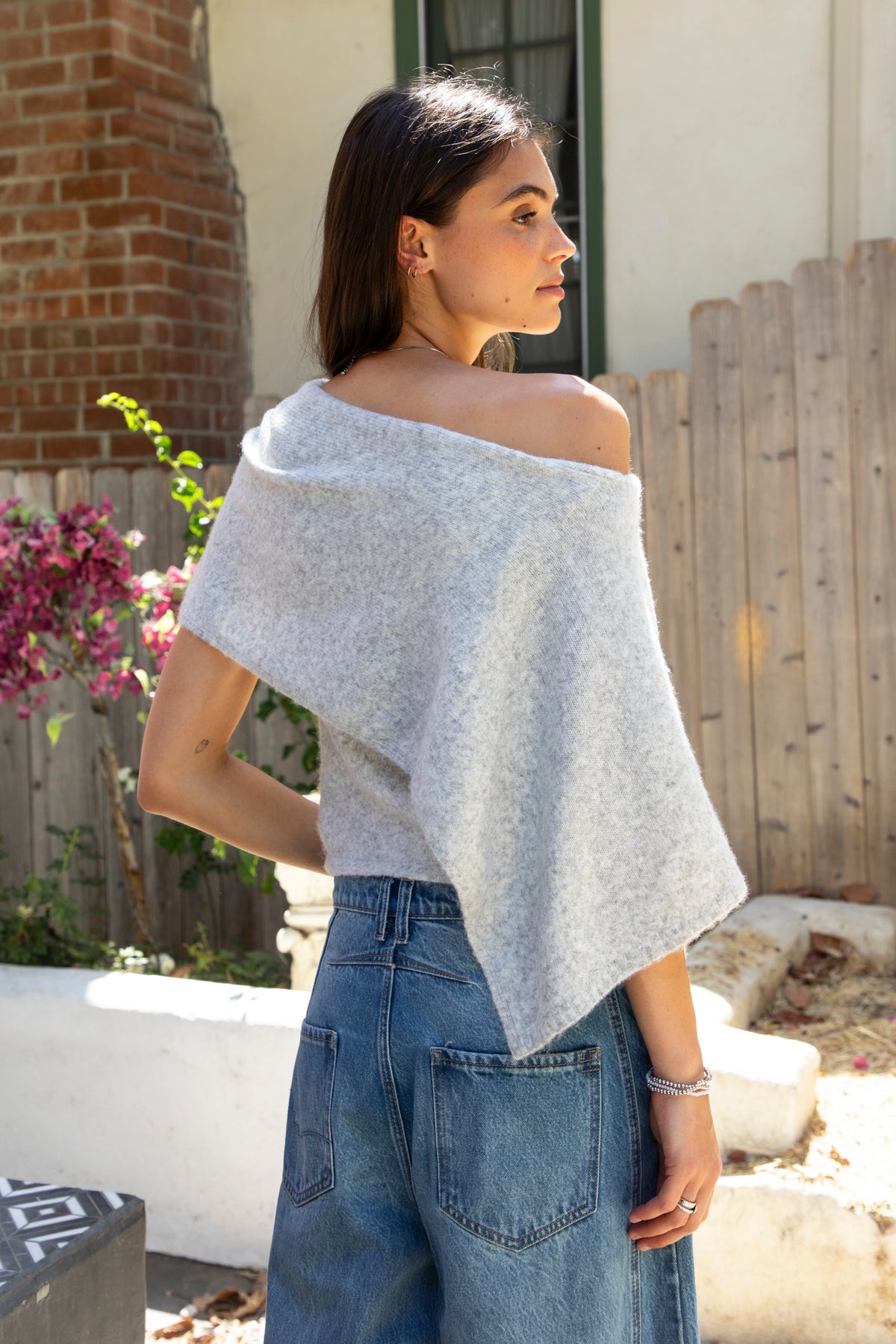 Woman wearing AINSLEY off shoulder asymmetric knit poncho sweater featuring soft brushed sweater knit fabric, cozy warmth, convertible off shoulder neckline, draped poncho silhouette, relaxed asymmetrical design, easy pull on style, and soft stretch comfort for fall and winter layering.