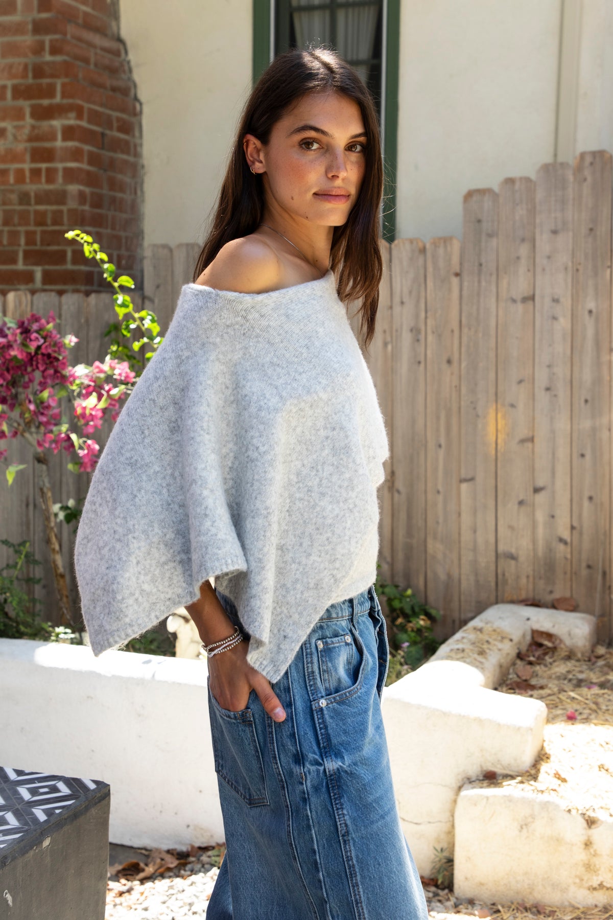 Woman wearing AINSLEY off shoulder asymmetric knit poncho sweater featuring soft brushed sweater knit fabric, cozy warmth, convertible off shoulder neckline, draped poncho silhouette, relaxed asymmetrical design, easy pull on style, and soft stretch comfort for fall and winter layering.