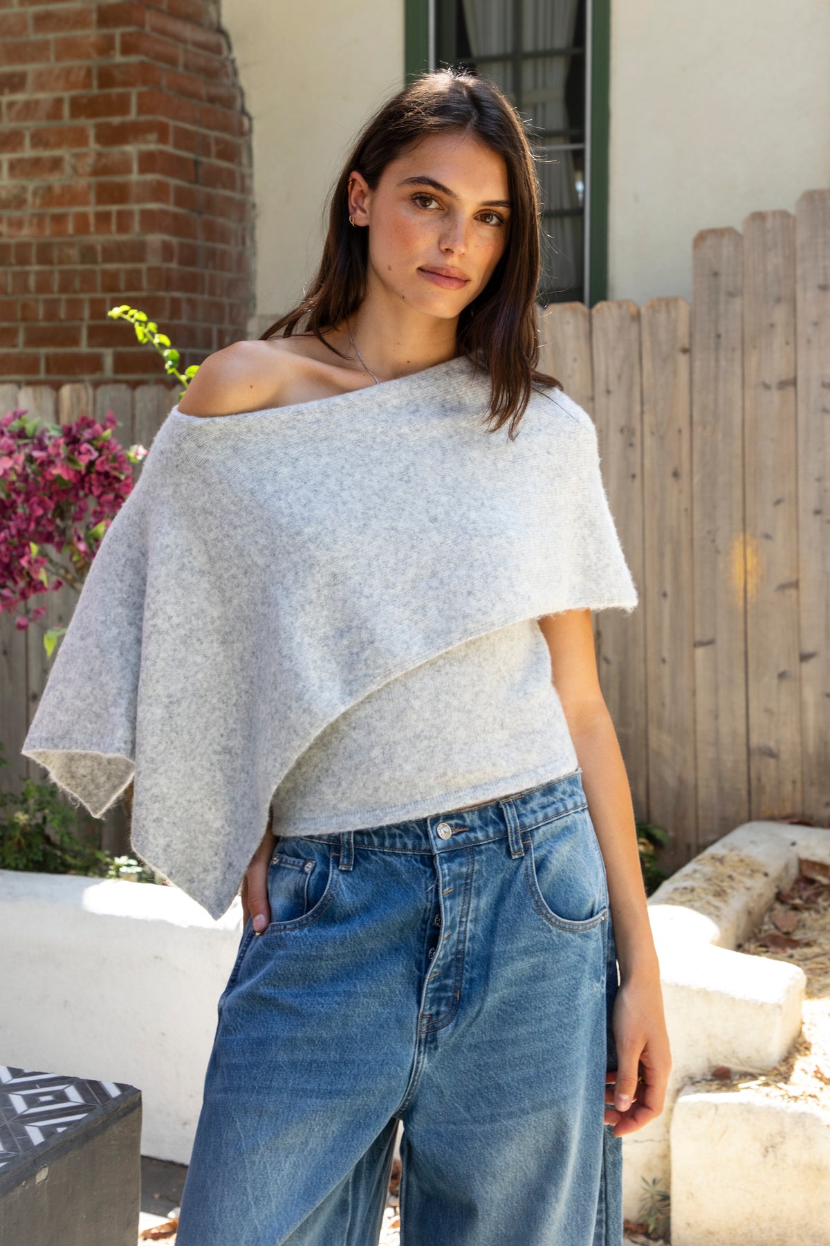 Woman wearing AINSLEY off shoulder asymmetric knit poncho sweater featuring soft brushed sweater knit fabric, cozy warmth, convertible off shoulder neckline, draped poncho silhouette, relaxed asymmetrical design, easy pull on style, and soft stretch comfort for fall and winter layering.