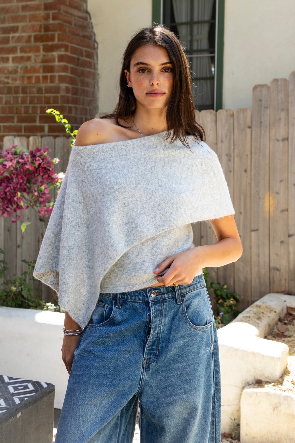 Woman wearing AINSLEY off shoulder asymmetric knit poncho sweater featuring soft brushed sweater knit fabric, cozy warmth, convertible off shoulder neckline, draped poncho silhouette, relaxed asymmetrical design, easy pull on style, and soft stretch comfort for fall and winter layering.