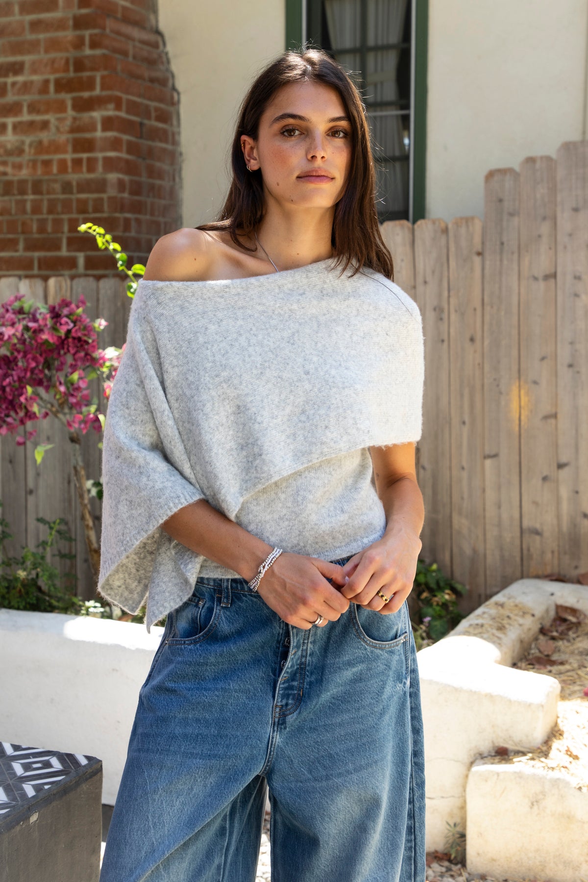 Woman wearing AINSLEY off shoulder asymmetric knit poncho sweater featuring soft brushed sweater knit fabric, cozy warmth, convertible off shoulder neckline, draped poncho silhouette, relaxed asymmetrical design, easy pull on style, and soft stretch comfort for fall and winter layering.
