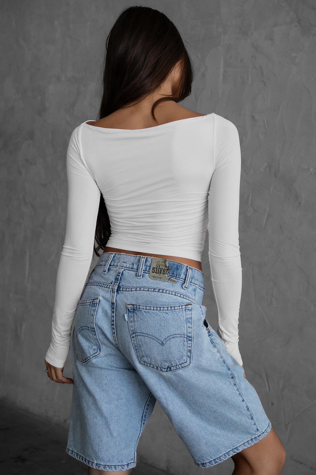 Women wearing TIC TOC JOLINA Lightweight Jersey Long Sleeve Top in white with ruched bodice, asymmetrical boat neckline, and cropped fit paired with blue denim agaisnt a wall. 