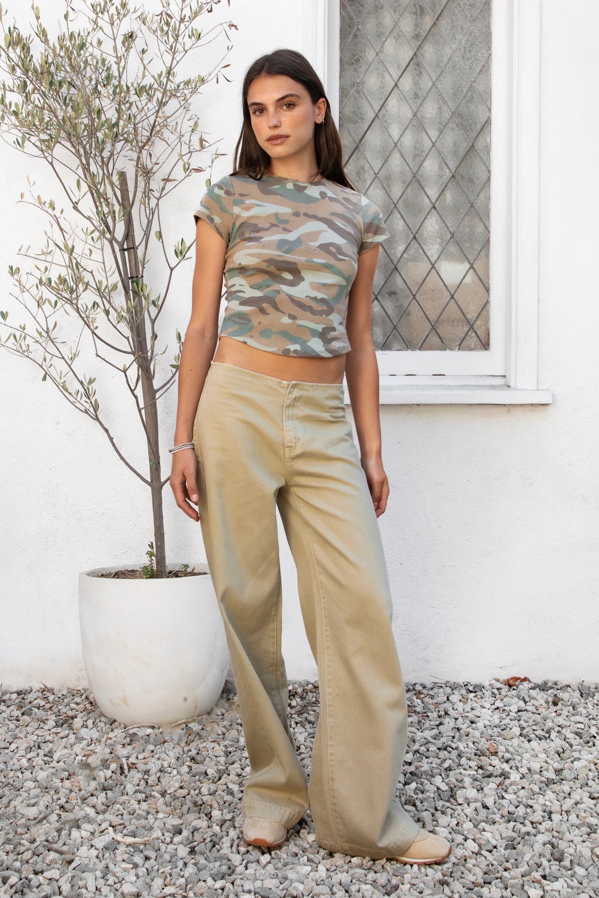 Woman wearing MAELYN military camo print fitted baby tee featuring soft single jersey fabric, comfortable stretch, allover camo print with muted color palette, classic crew neckline, short sleeves, fitted body skimming shape, and slightly cropped length that sits naturally at the waist.