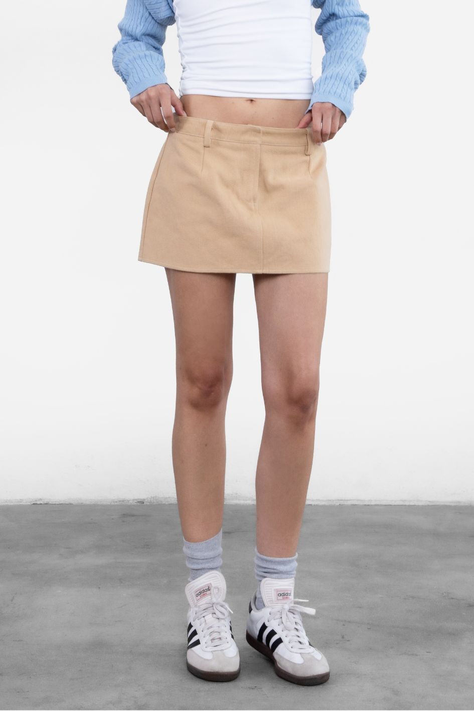 Person wearing a beige skirt, light blue sweater, white shirt, Adidas sneakers, and gray socks on a plain background.