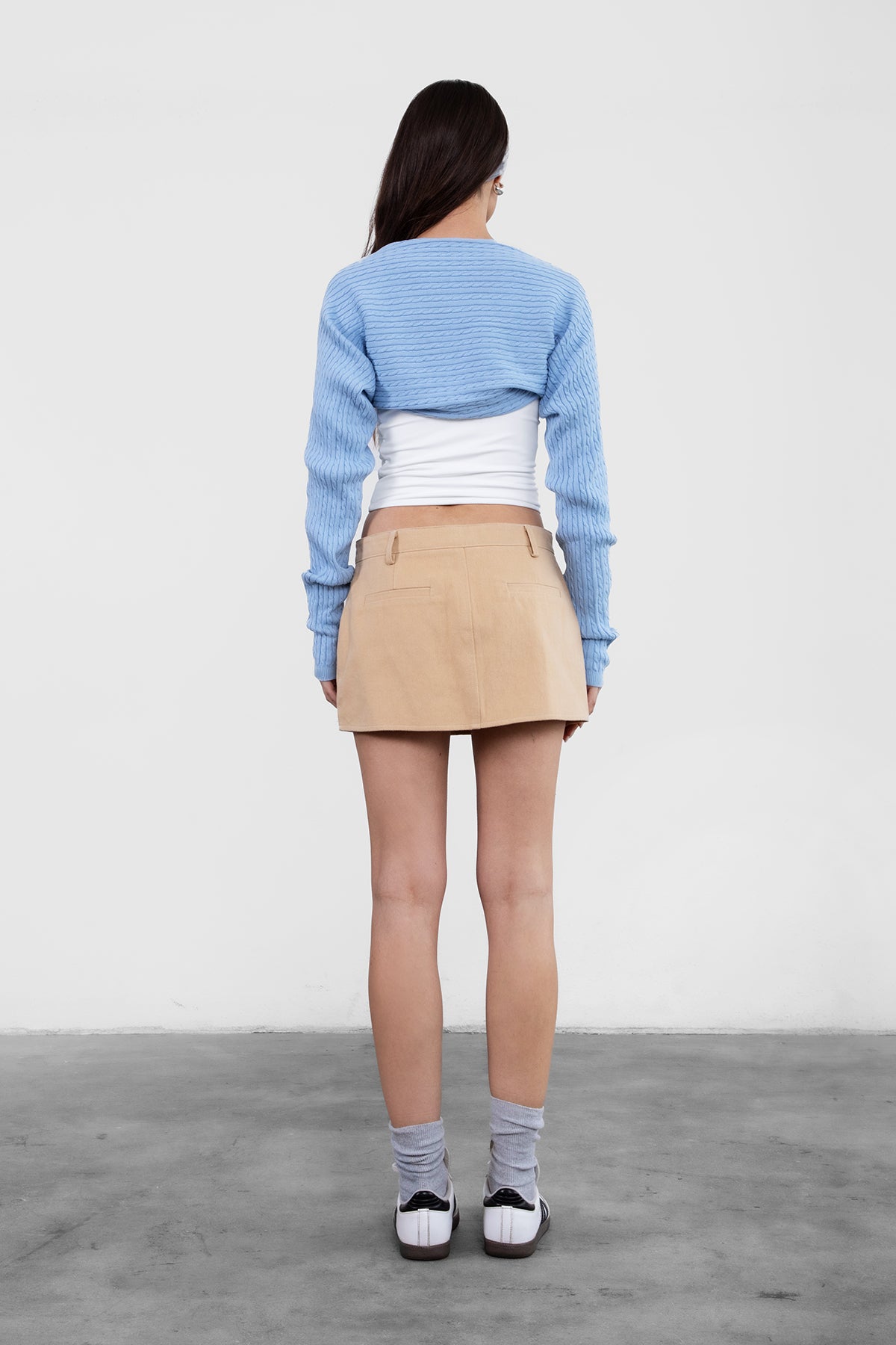 Person wearing a blue sweater, white top, beige skirt, and black shoes on a plain background
