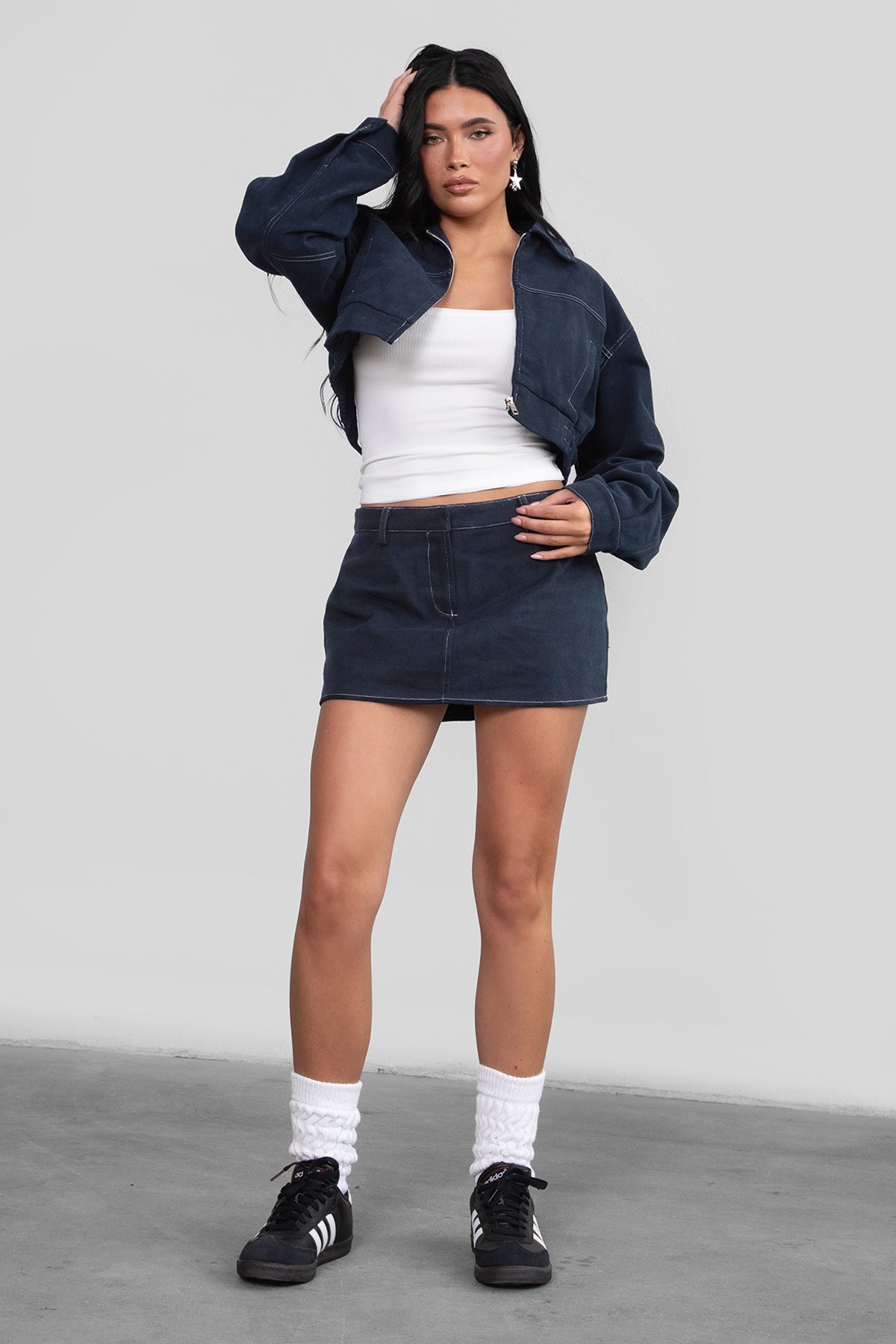 Woman wearing a navy jacket, white top, and denim skirt against a plain background