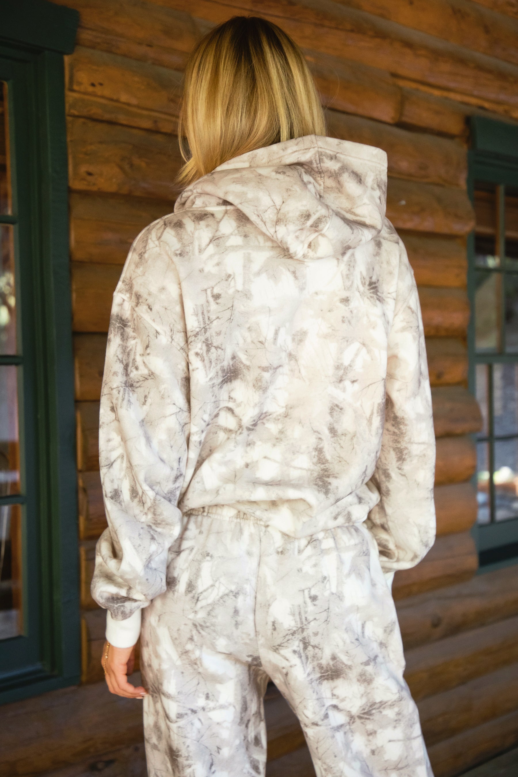 Woman wearing a TIC TOC AVERY Woodland Camo Oversized Hoodie with kangaroo pocket, ribbed trim, and soft Cloud Fleece fabric, standing in front of a wooden wall.