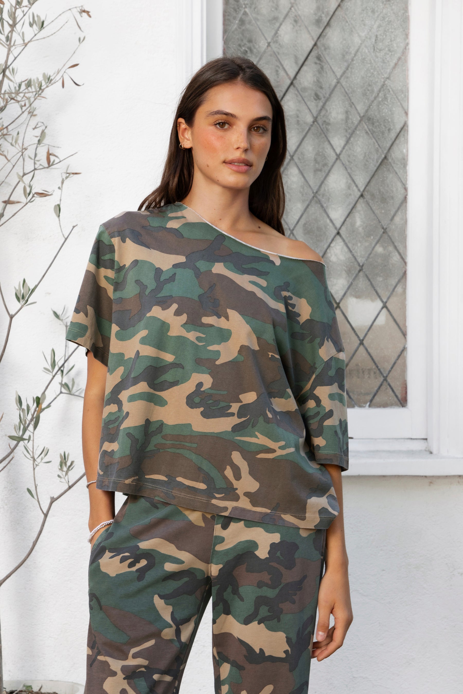 Woman wearing SCOUT military camo short sleeve oversized off shoulder top featuring soft breathable single jersey fabric, allover classic camouflage print, wide rolled raw edge neckline, relaxed oversized silhouette, dropped shoulders, elbow length sleeves, and casual boyfriend inspired draped fit.