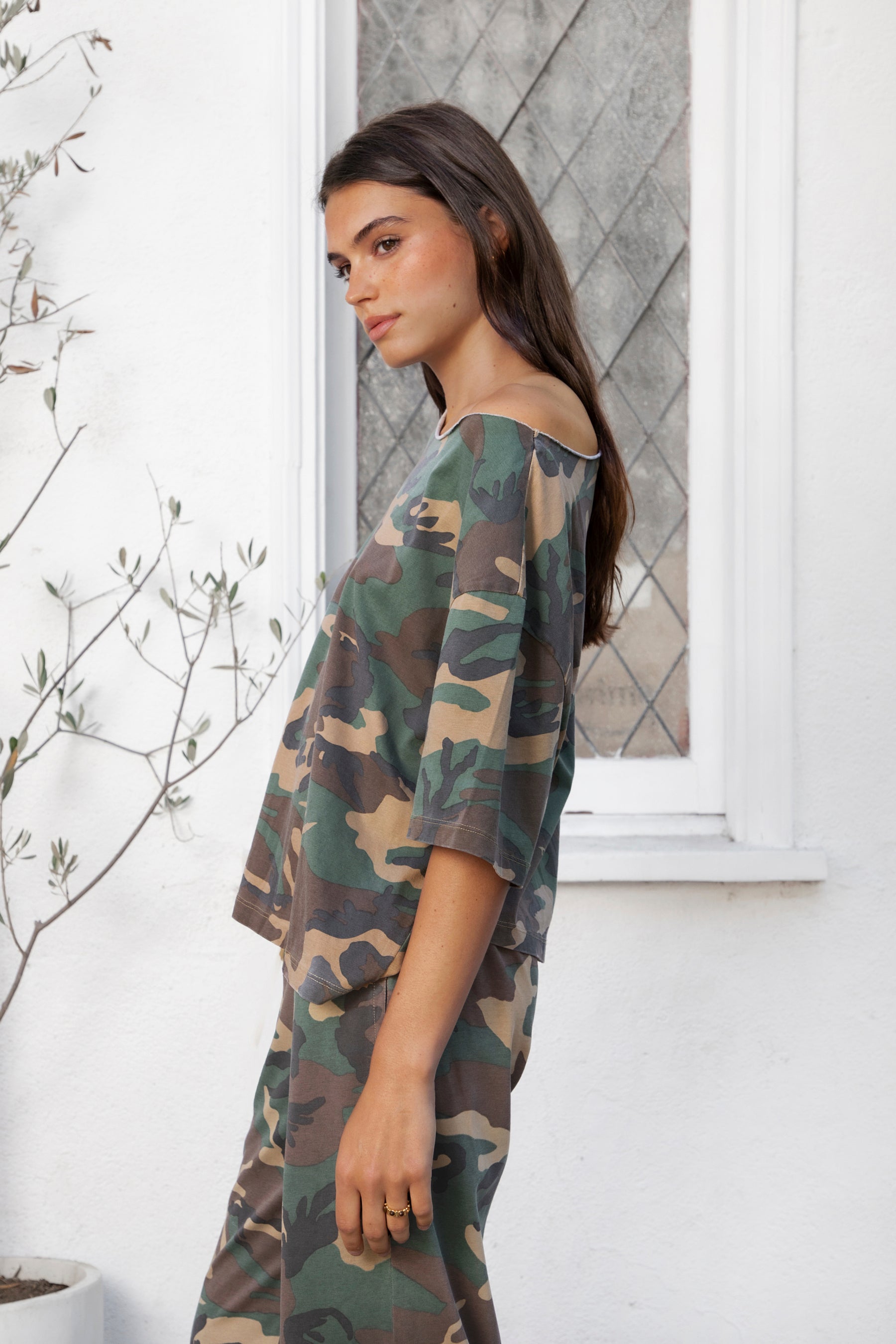 Woman wearing SCOUT military camo short sleeve oversized off shoulder top featuring soft breathable single jersey fabric, allover classic camouflage print, wide rolled raw edge neckline, relaxed oversized silhouette, dropped shoulders, elbow length sleeves, and casual boyfriend inspired draped fit.