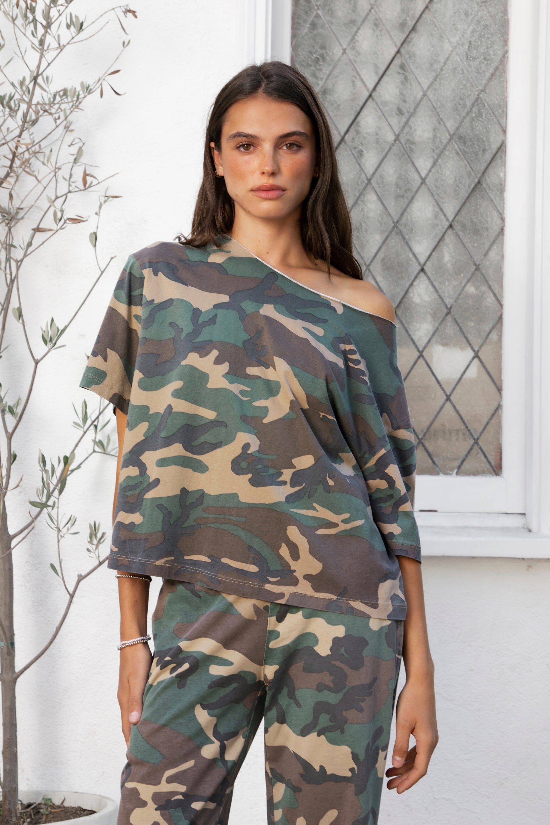 Woman wearing SCOUT military camo short sleeve oversized off shoulder top featuring soft breathable single jersey fabric, allover classic camouflage print, wide rolled raw edge neckline, relaxed oversized silhouette, dropped shoulders, elbow length sleeves, and casual boyfriend inspired draped fit.