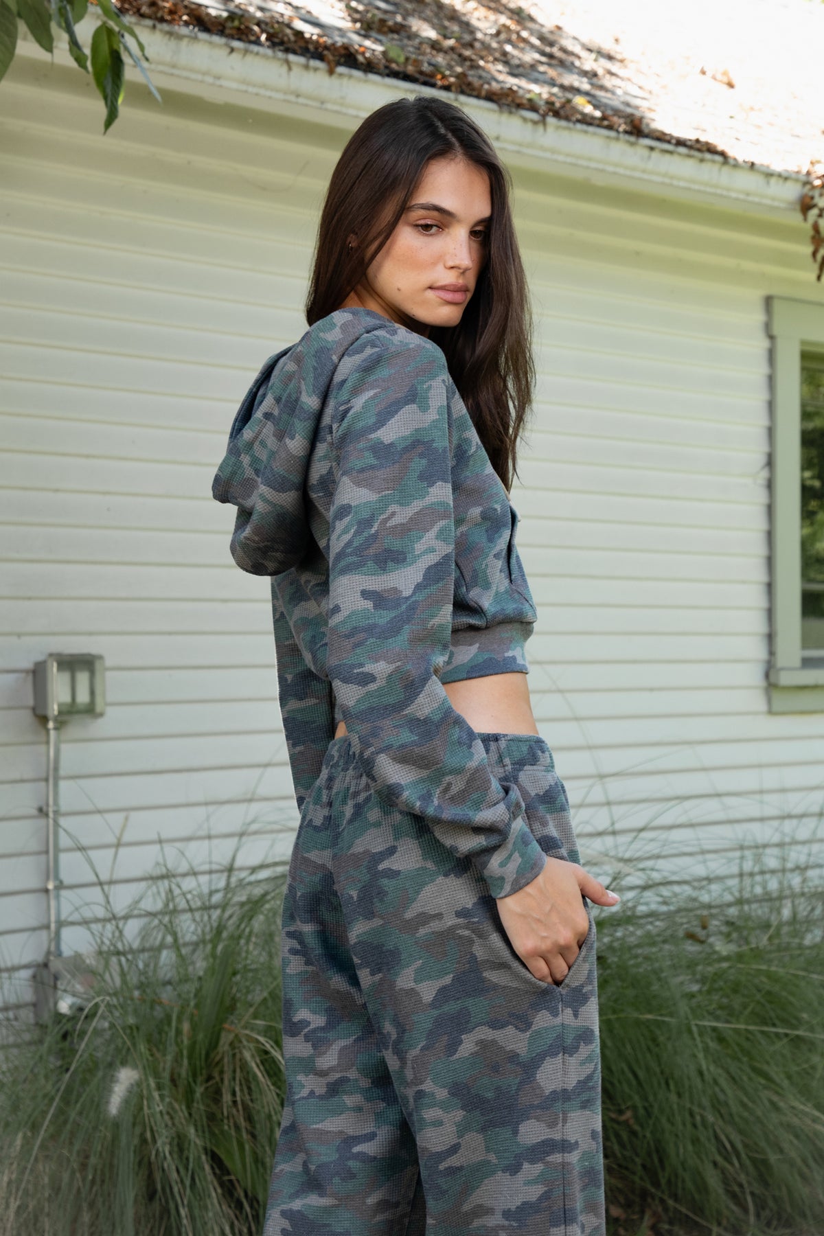 Woman wearing AMAIA heavy weight military camo zip up hoodie featuring heavyweight waffle knit fabric with a soft textured feel, allover woodland camouflage print in muted green and grey tones, relaxed cropped silhouette, attached hood with adjustable drawstrings, full front zipper closure, long sleeves with ribbed cuffs, functional front kangaroo pockets, and wide ribbed waistband.