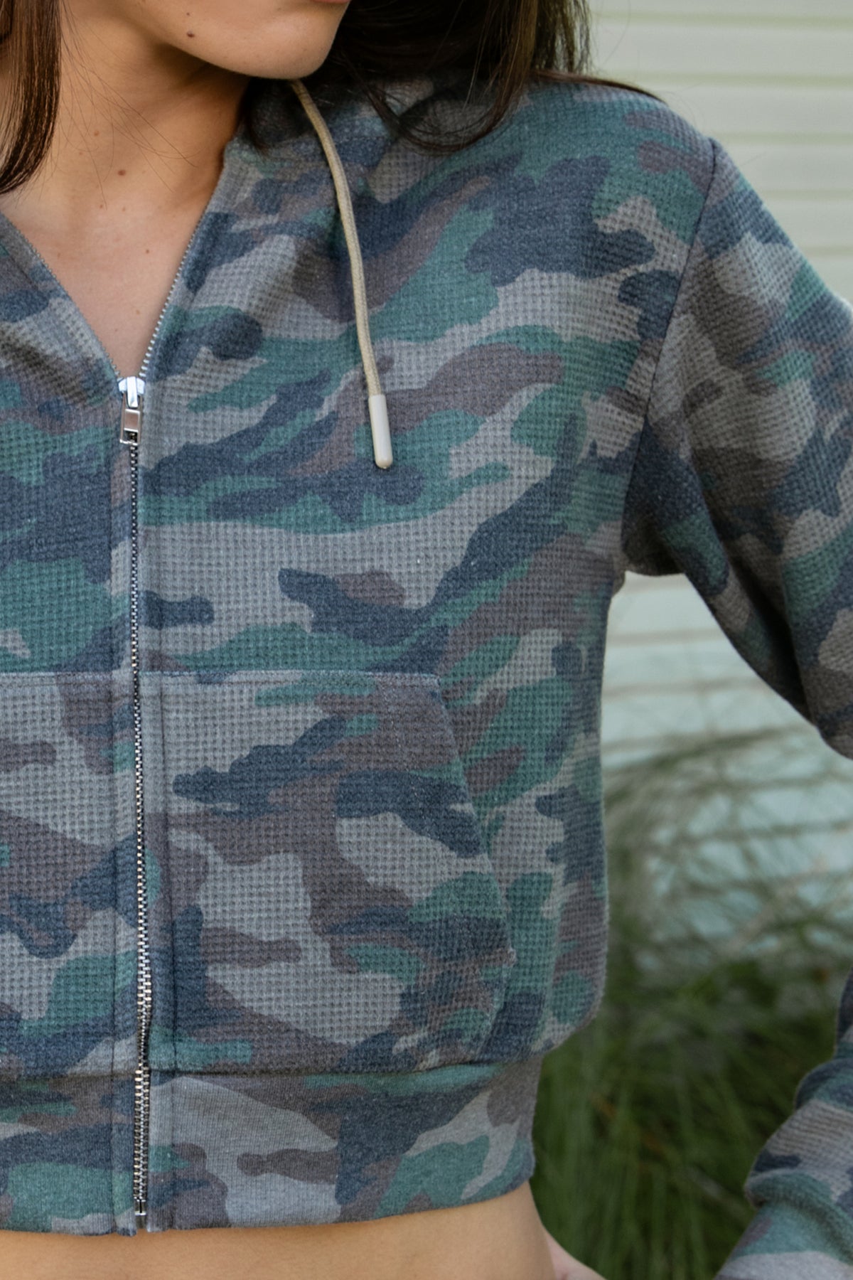 Woman wearing AMAIA heavy weight military camo zip up hoodie featuring heavyweight waffle knit fabric with a soft textured feel, allover woodland camouflage print in muted green and grey tones, relaxed cropped silhouette, attached hood with adjustable drawstrings, full front zipper closure, long sleeves with ribbed cuffs, functional front kangaroo pockets, and wide ribbed waistband.