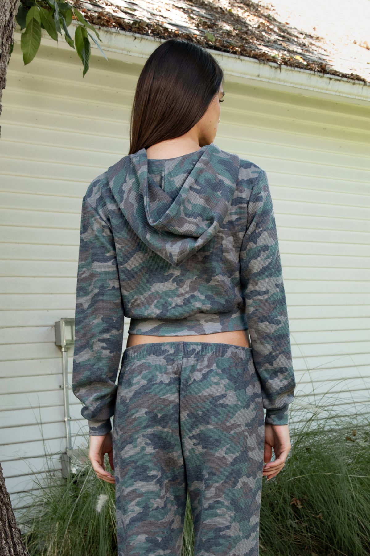 Woman wearing AMAIA heavy weight military camo zip up hoodie featuring heavyweight waffle knit fabric with a soft textured feel, allover woodland camouflage print in muted green and grey tones, relaxed cropped silhouette, attached hood with adjustable drawstrings, full front zipper closure, long sleeves with ribbed cuffs, functional front kangaroo pockets, and wide ribbed waistband.