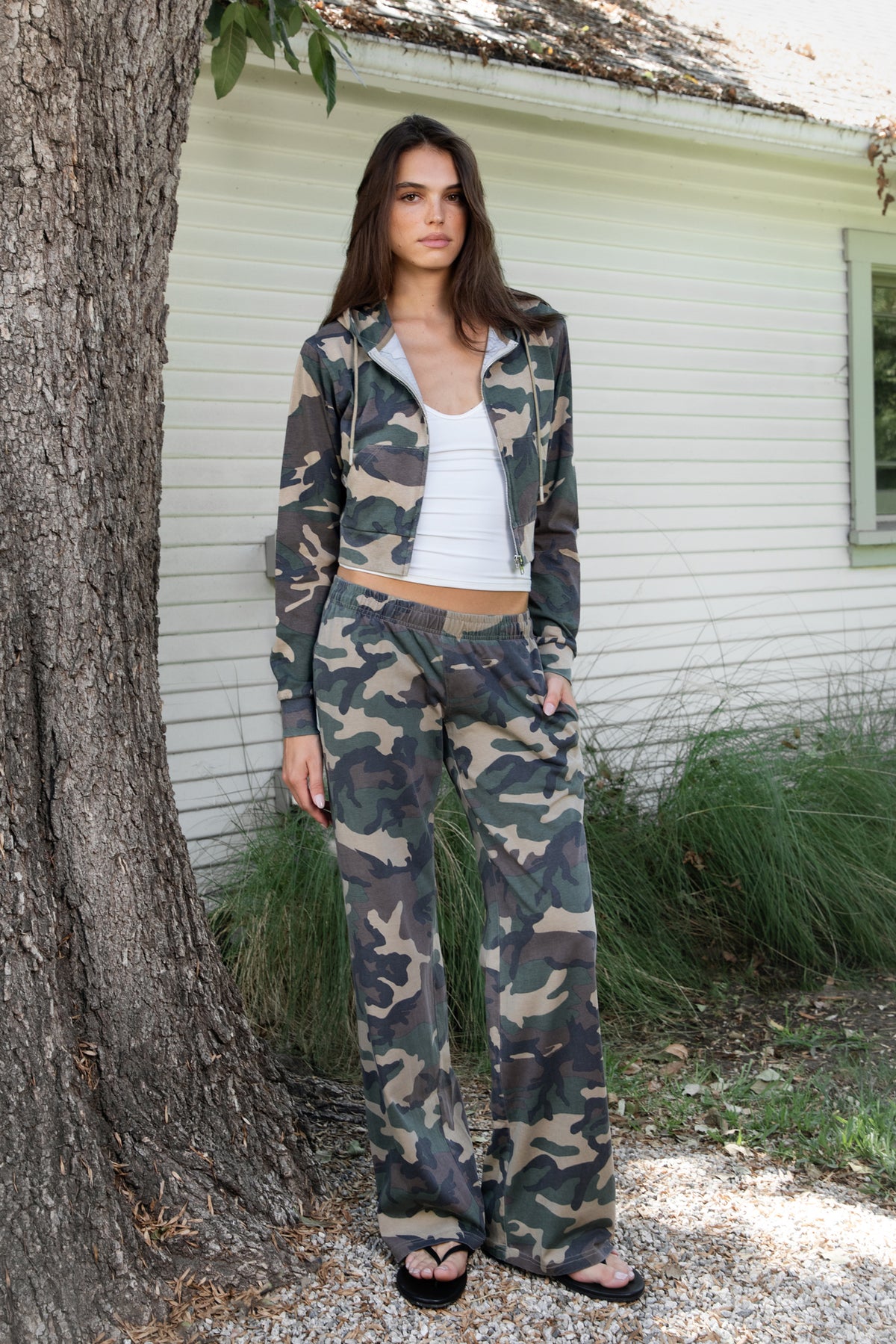 Woman wearing SCOUT military camo crop zip up hoodie featuring soft cotton single jersey fabric, allover woodland camouflage print in earthy green and brown tones, relaxed cropped silhouette, attached hood with adjustable drawstrings, full front zipper closure, long sleeves with ribbed cuffs, functional front kangaroo pockets, and ribbed waistband for a comfortable finish.