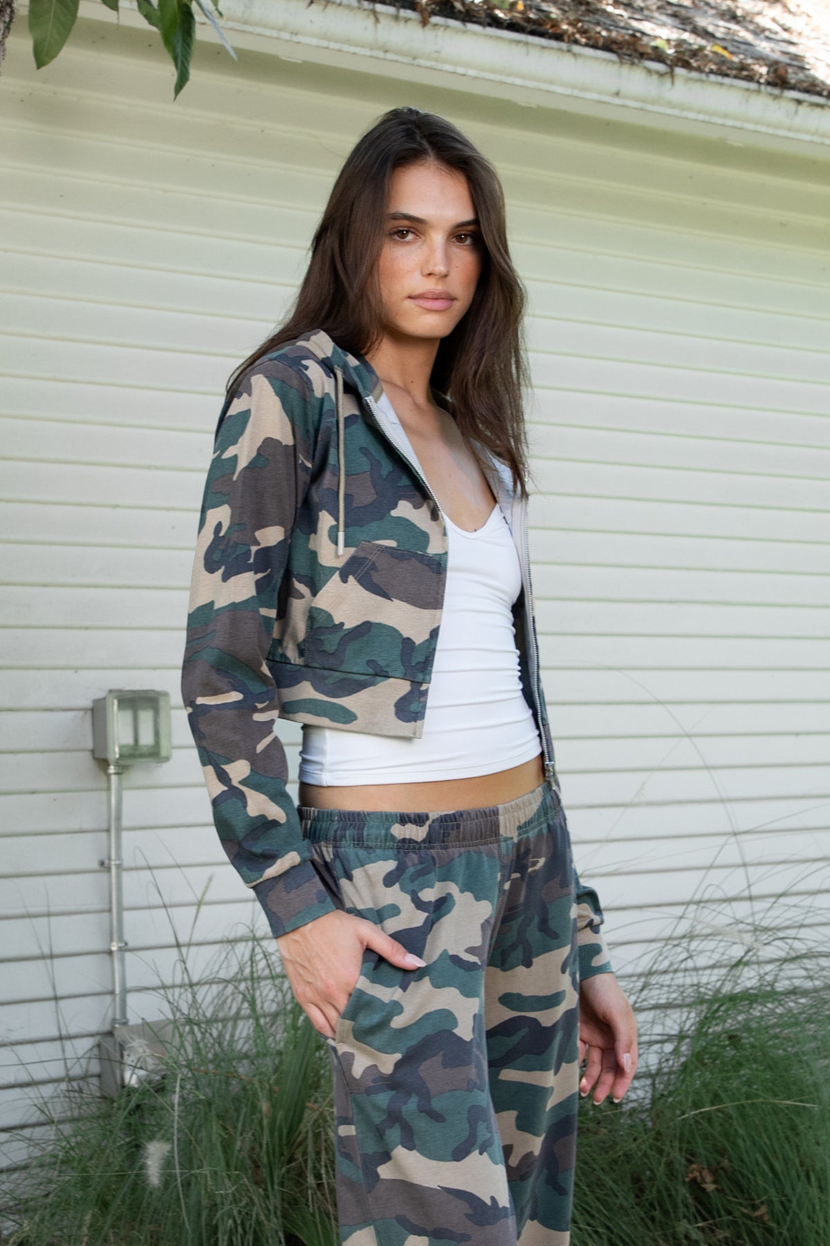 Woman wearing SCOUT military camo crop zip up hoodie featuring soft cotton single jersey fabric, allover woodland camouflage print in earthy green and brown tones, relaxed cropped silhouette, attached hood with adjustable drawstrings, full front zipper closure, long sleeves with ribbed cuffs, functional front kangaroo pockets, and ribbed waistband for a comfortable finish.
