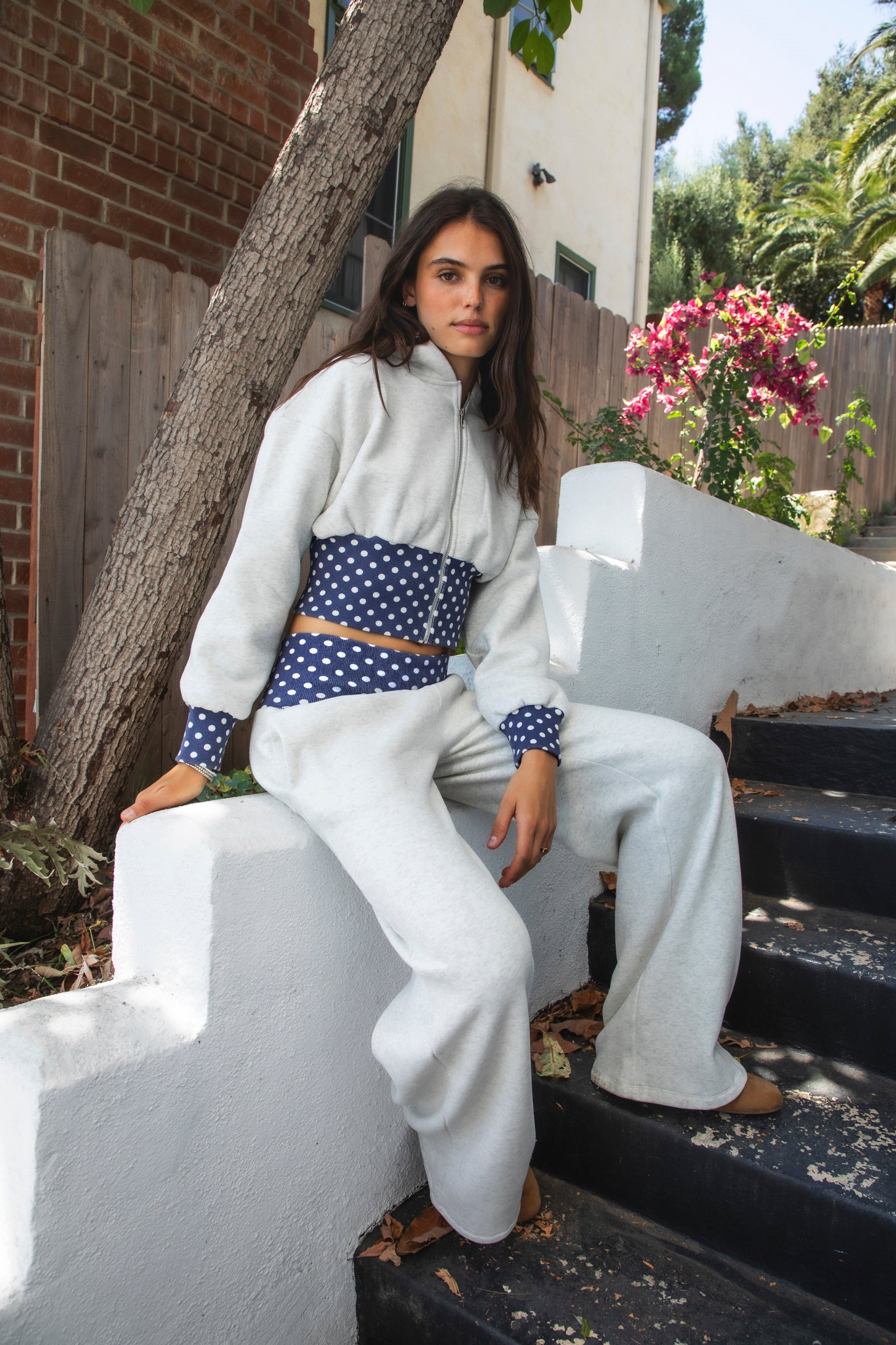 Woman wearing COSETTE polka dot corset waist cropped zip up hoodie featuring soft fleece fabric, contrast navy ribbed corset inspired waistband with allover white polka dot pattern, fitted extended waist, matching polka dot cuffs, full front zipper closure, relaxed drop shoulder sleeves, attached hood, and cropped length designed to coordinate with the matching pants.
