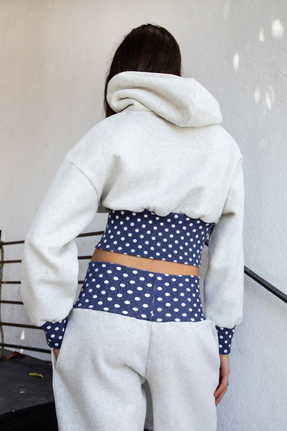 Woman wearing COSETTE polka dot corset waist cropped zip up hoodie featuring soft fleece fabric, contrast navy ribbed corset inspired waistband with allover white polka dot pattern, fitted extended waist, matching polka dot cuffs, full front zipper closure, relaxed drop shoulder sleeves, attached hood, and cropped length designed to coordinate with the matching pants.