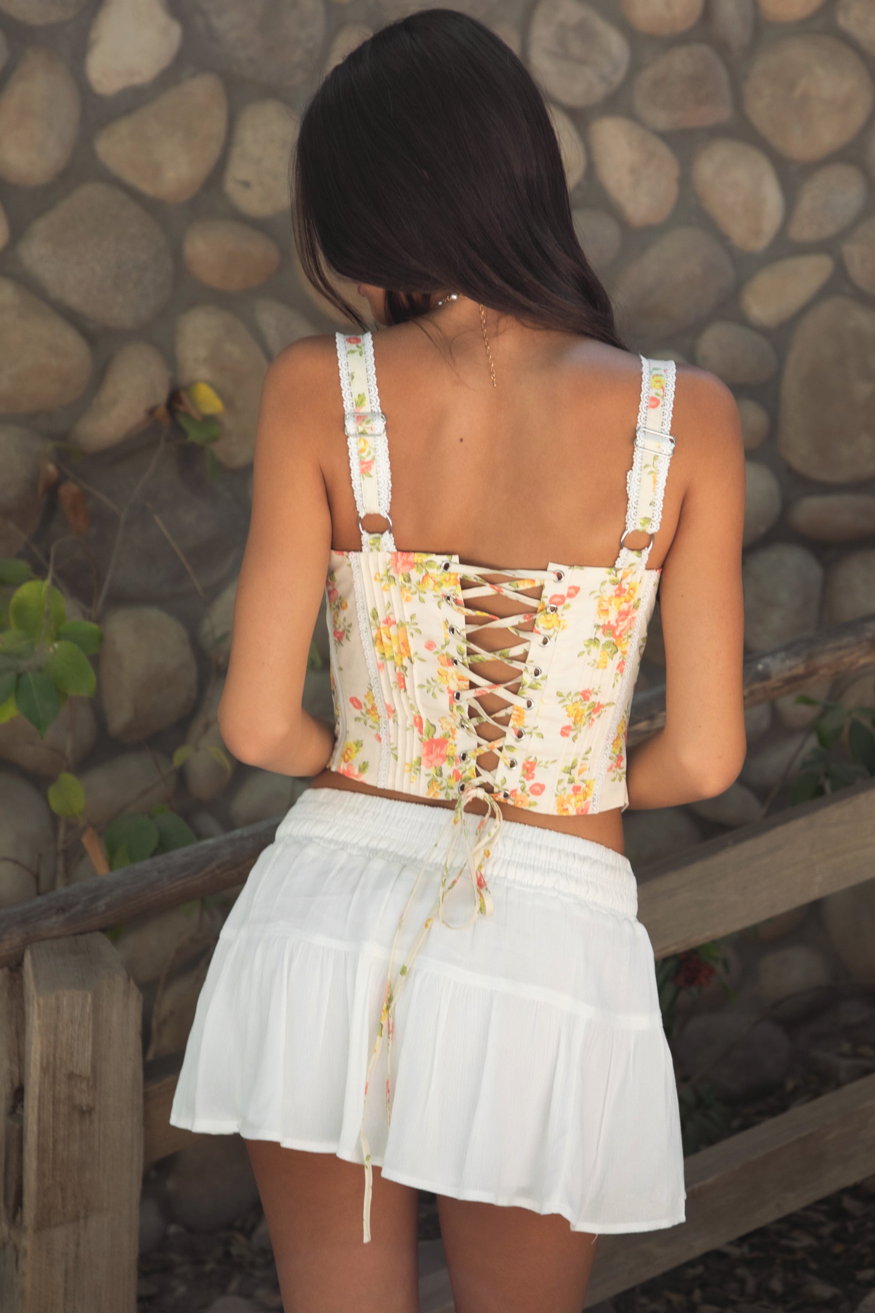 Woman wearing TIC TOC ELORA Floral Lace Bustier Top, back view showcasing lace-up detail, paired with a white tiered mini skirt, outdoors. 