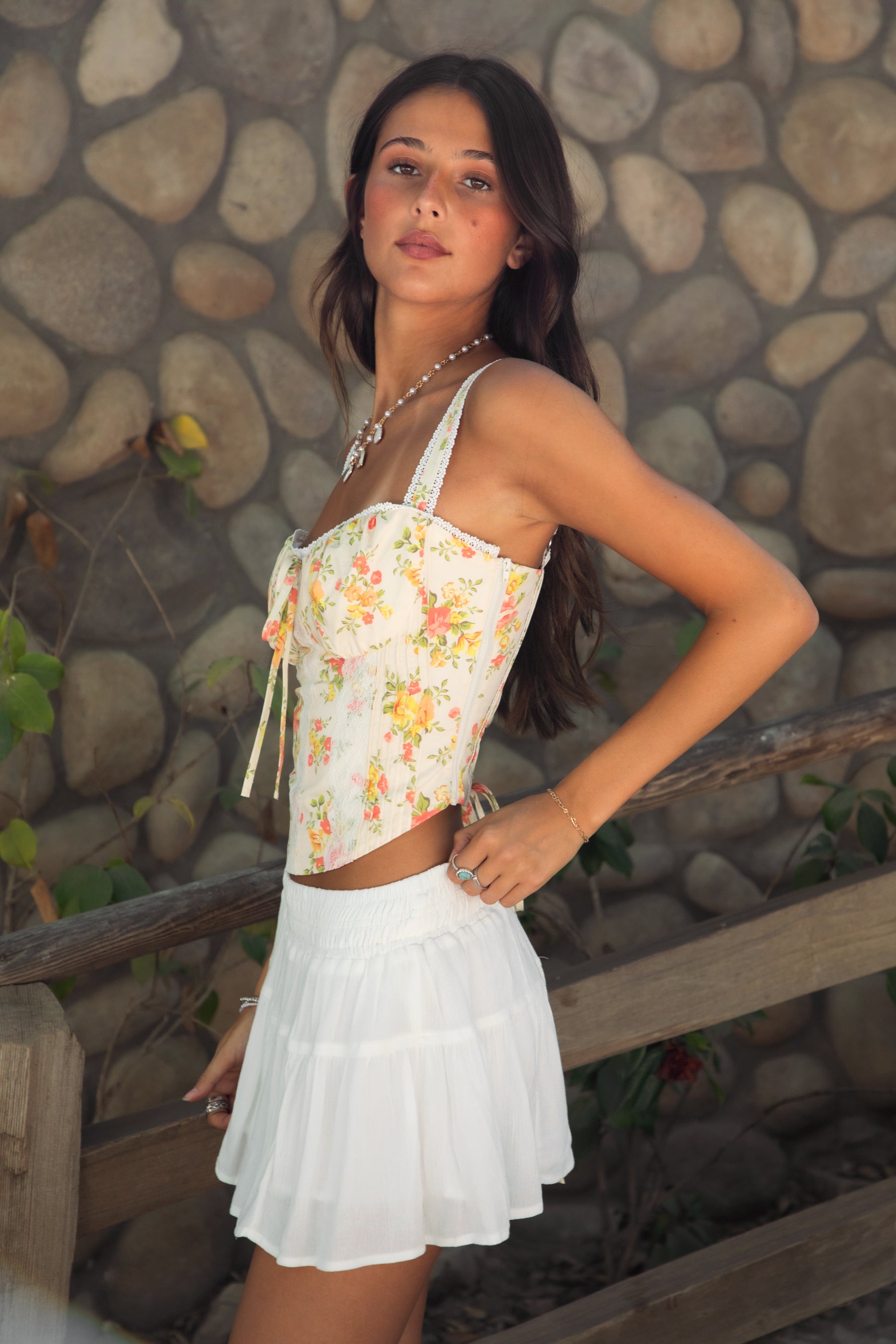Woman wearing TIC TOC ELORA Floral Lace Bustier Top, side view showing the sweetheart neckline and side zipper closure, paired with a white tiered mini skirt, outdoors. 