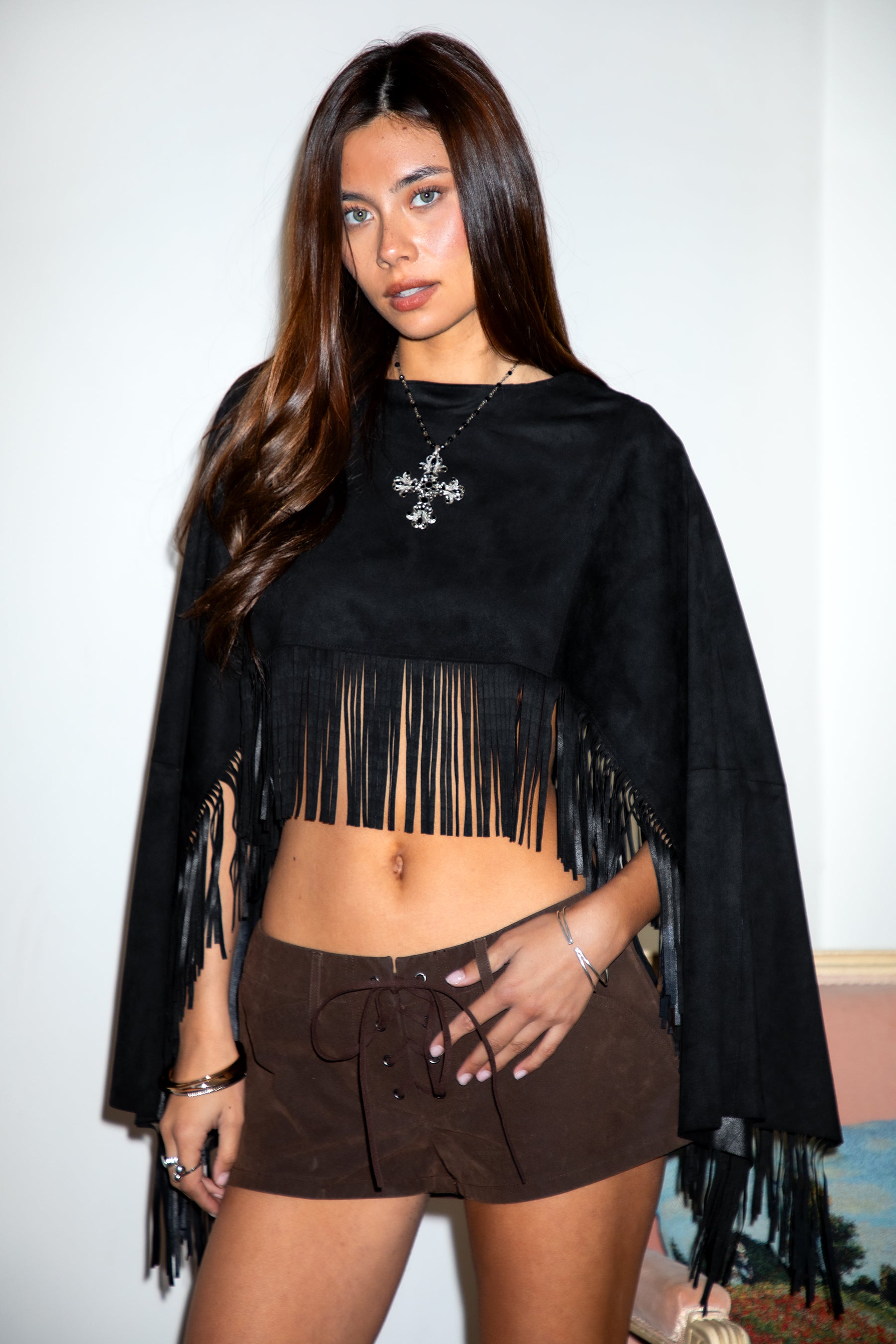 Woman wearing TIC TOC TL31825 STALLION faux suede fringe poncho featuring a cropped cape silhouette, drop shoulder relaxed fit, long fringe tassel detailing along the hem and sleeves, and a soft western-inspired faux suede texture, styled over a fitted top with denim bottoms for a rodeo festival look.
