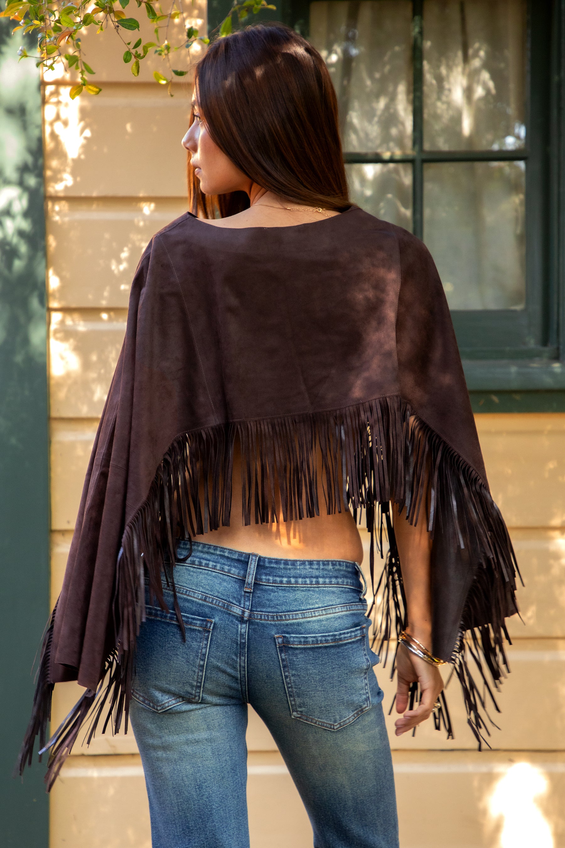 Woman wearing TIC TOC TL31825 STALLION faux suede fringe poncho featuring a cropped cape silhouette, drop shoulder relaxed fit, long fringe tassel detailing along the hem and sleeves, and a soft western-inspired faux suede texture, styled over a fitted top with denim bottoms for a rodeo festival look.