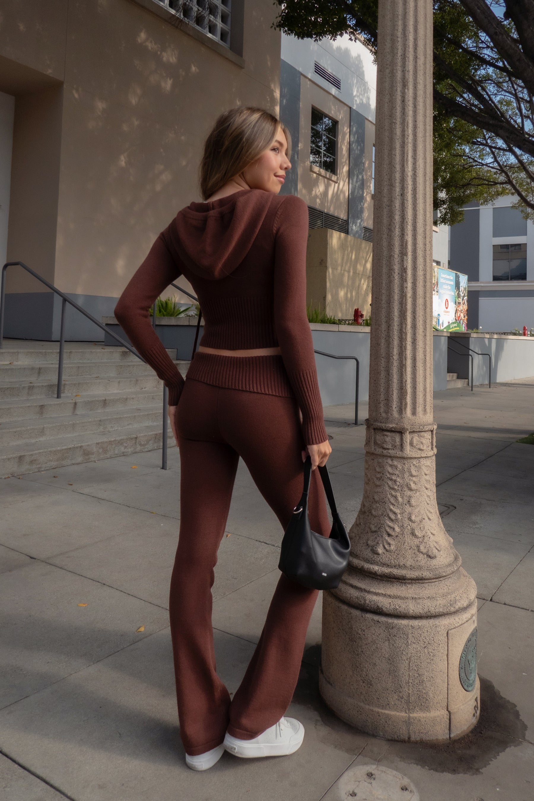 Woman wearing TIC TOC TPC29297-COZEE brown sweater knit two-piece set featuring a cropped zip-up hoodie with ribbed details and matching fold-over flare pants, styled outdoors with sneakers and a small black shoulder bag.