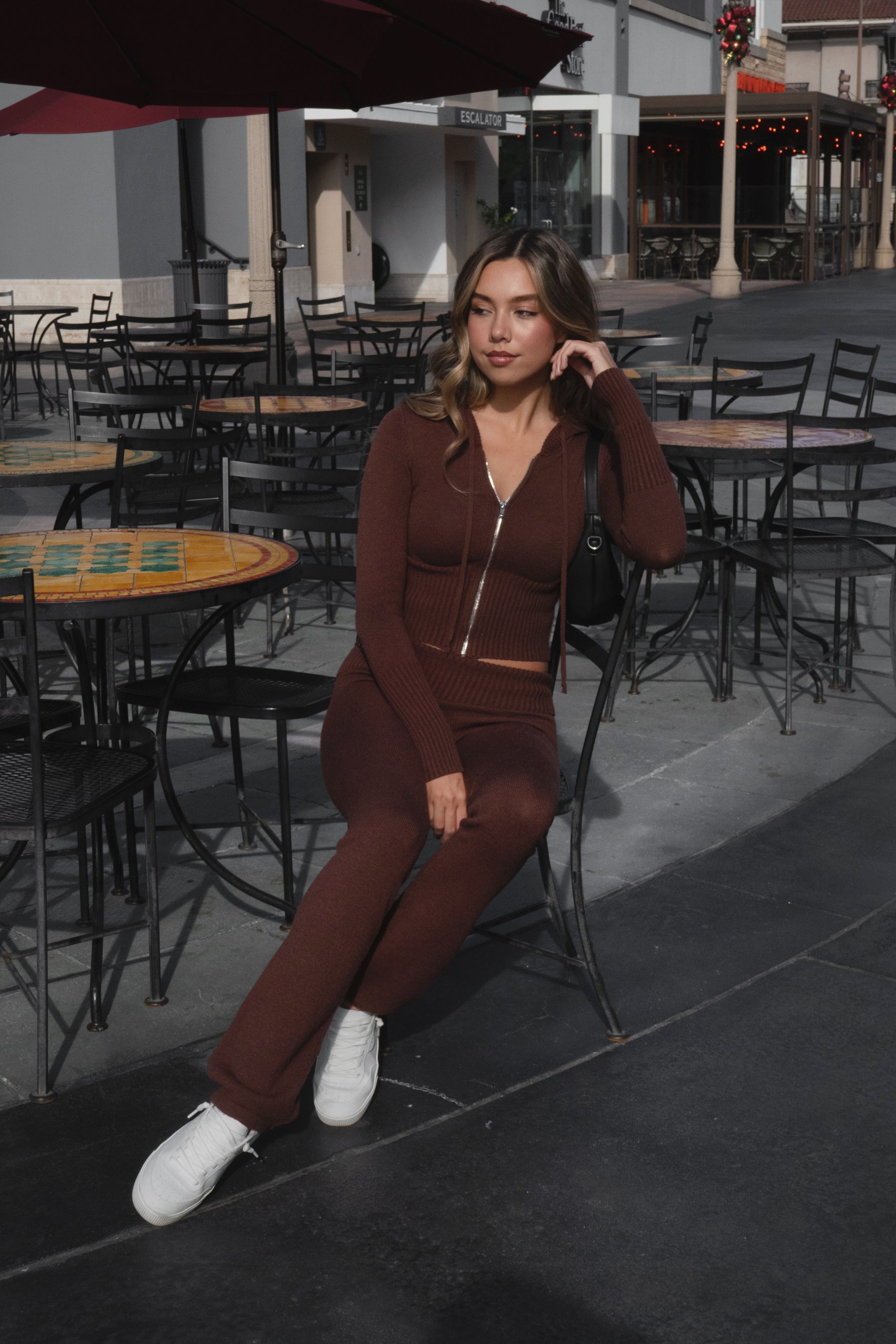 Woman wearing TIC TOC TPC29297-COZEE brown sweater knit two-piece set featuring a cropped zip-up hoodie with ribbed details and matching fold-over flare pants, styled outdoors with sneakers and a small black shoulder bag.
