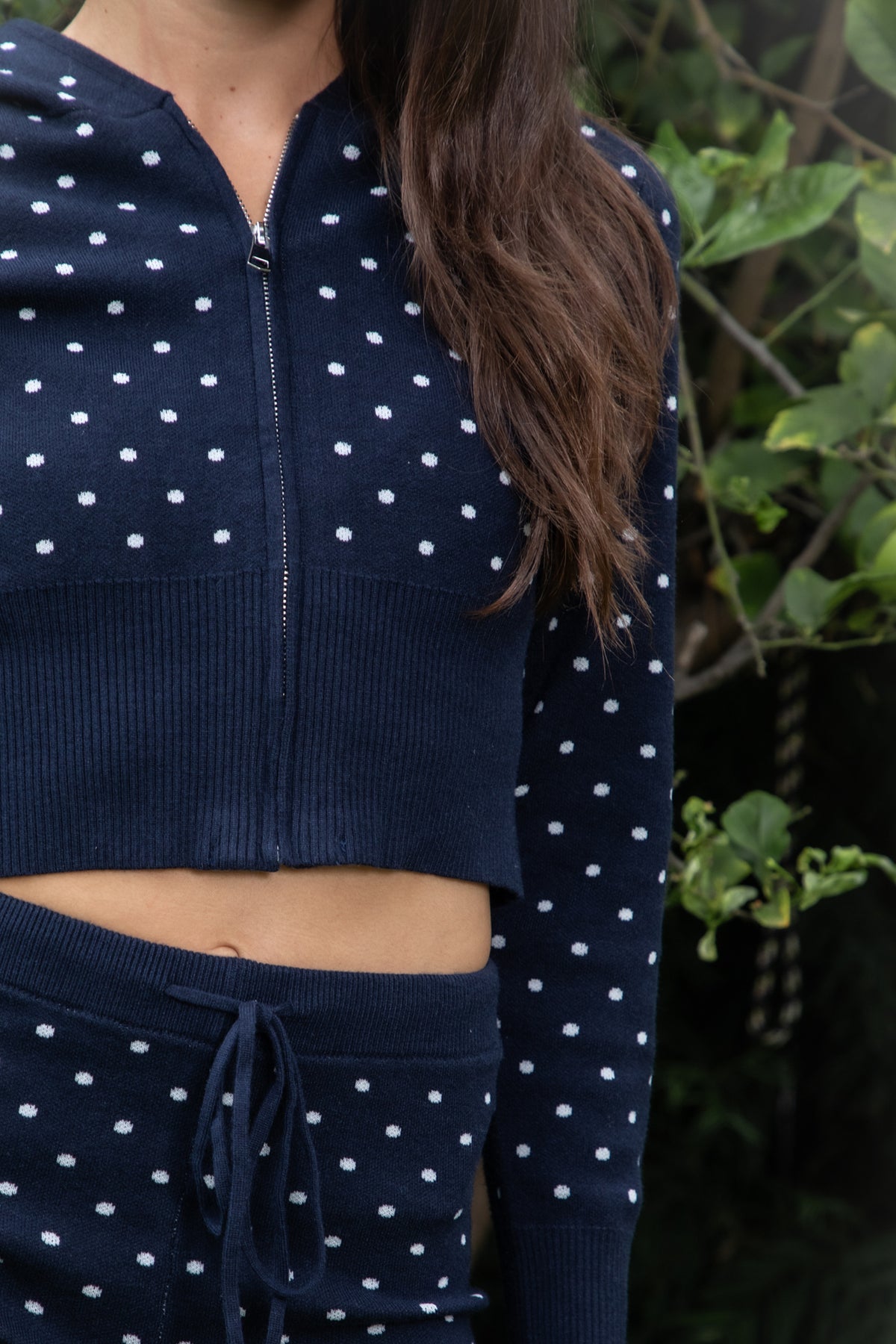 Woman wearing DOTELLE polka dot knit hoodie and shorts set featuring soft stretch sweater knit fabric with an allover polka dot pattern, cropped zip up hoodie with adjustable drawstring hood, fitted long sleeves with ribbed cuffs, wide ribbed waistband and hem, and matching high waisted fitted shorts with adjustable drawstring waist and short length.