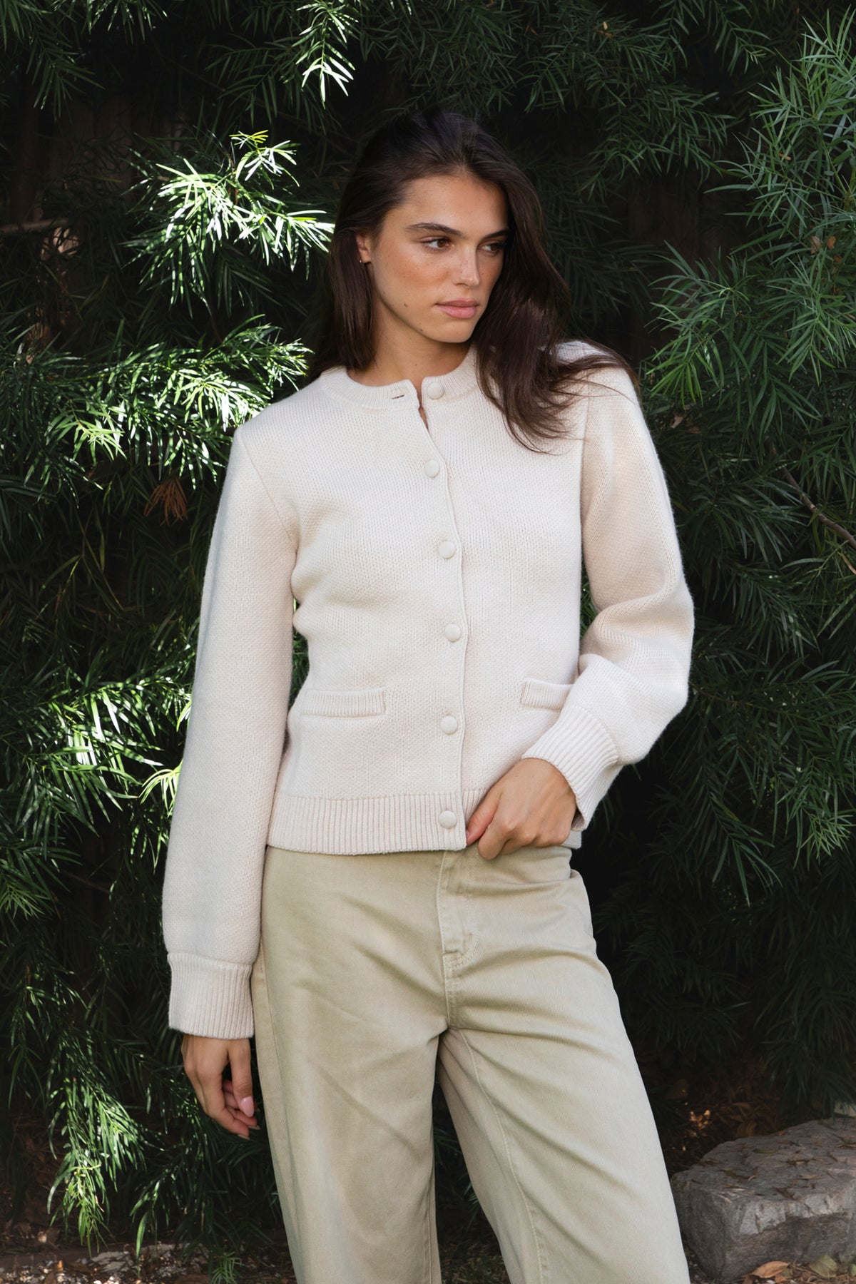 Woman wearing SABINE button front knit cardigan featuring soft textured sweater knit fabric with anti pilling finish, classic crew neckline with ribbed trim, full front closure with matching fabric covered buttons, long sleeves with ribbed cuffs, decorative front welt pockets, relaxed straight fit, slightly fitted waist, ribbed hem, and hip length silhouette.