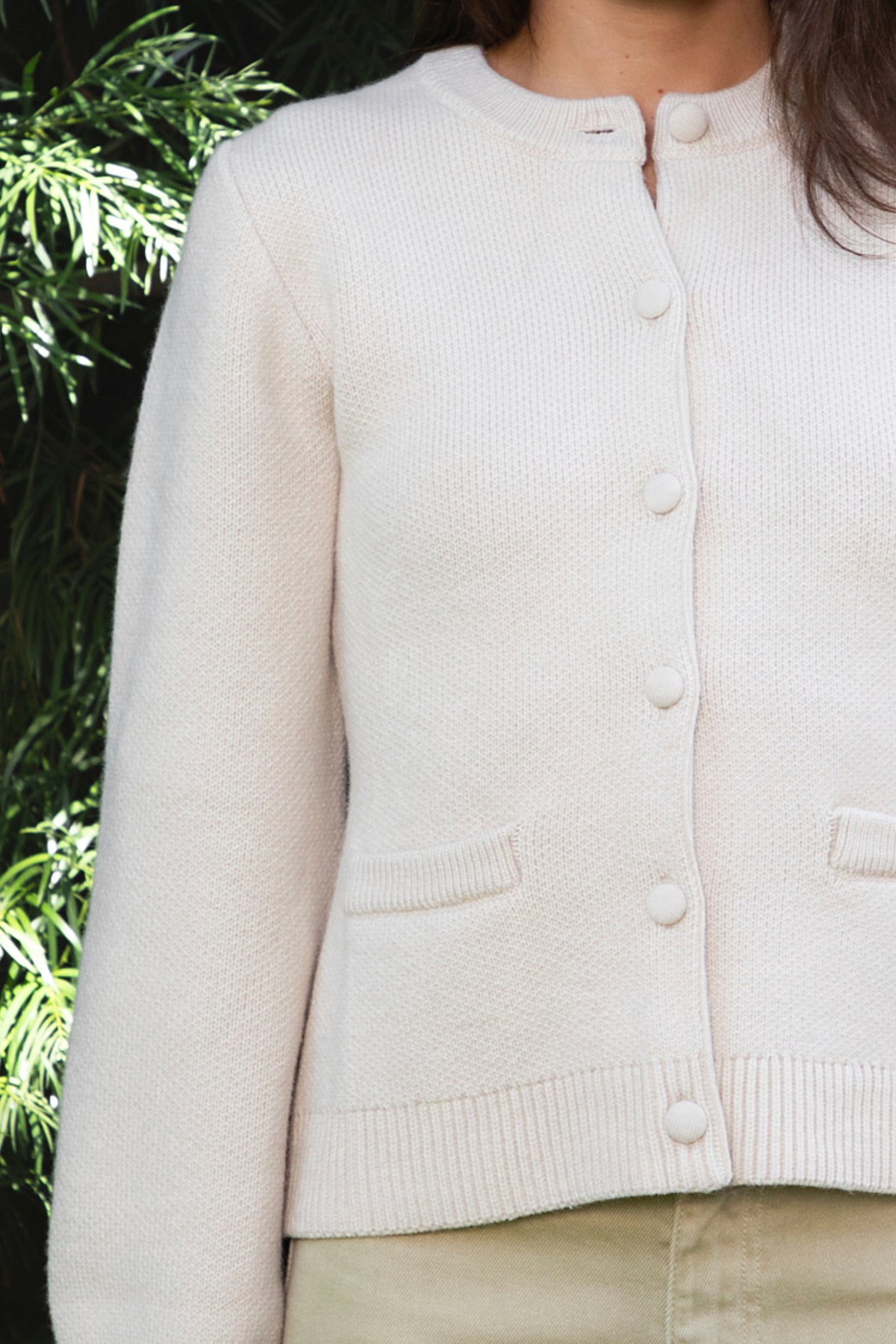Woman wearing SABINE button front knit cardigan featuring soft textured sweater knit fabric with anti pilling finish, classic crew neckline with ribbed trim, full front closure with matching fabric covered buttons, long sleeves with ribbed cuffs, decorative front welt pockets, relaxed straight fit, slightly fitted waist, ribbed hem, and hip length silhouette.