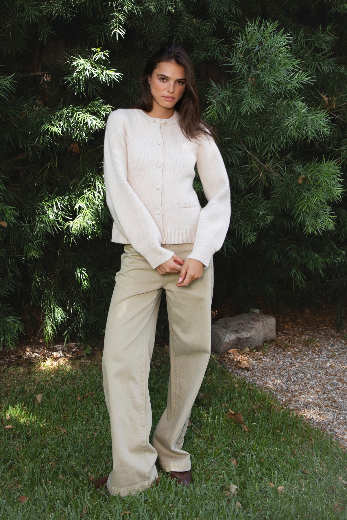 Woman wearing SABINE button front knit cardigan featuring soft textured sweater knit fabric with anti pilling finish, classic crew neckline with ribbed trim, full front closure with matching fabric covered buttons, long sleeves with ribbed cuffs, decorative front welt pockets, relaxed straight fit, slightly fitted waist, ribbed hem, and hip length silhouette.