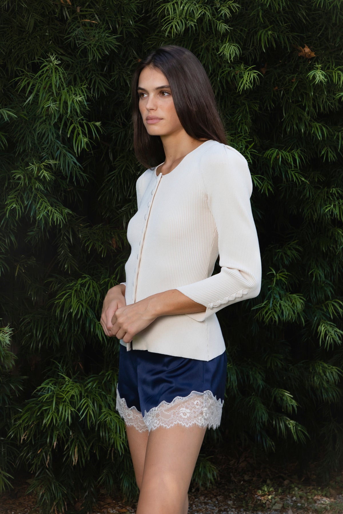 Woman wearing VIVIENNE lace up ribbed knit cardigan featuring fitted ribbed sweater knit fabric, soft scoop neckline, covered button front closure, long sleeves with subtle puff shoulder shaping, lace up detailing along the sleeve cuffs, decorative lace up detail down the center back, and slightly curved hem with fitted waist shaping.