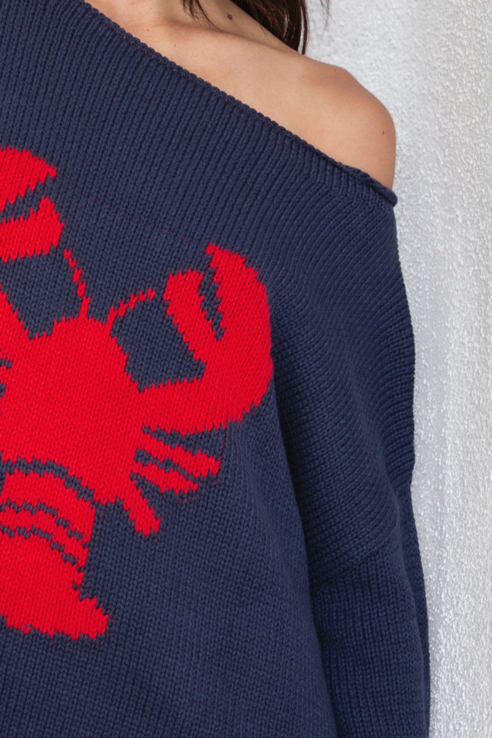 Woman wearing LOBELLE lobster graphic off shoulder sweater featuring relaxed sweater knit fabric, bold contrast red lobster intarsia graphic at the front, wide neckline designed to drape off the shoulder, oversized slouchy silhouette, dropped shoulders, long relaxed sleeves, ribbed cuffs and hem, and subtle side slits.