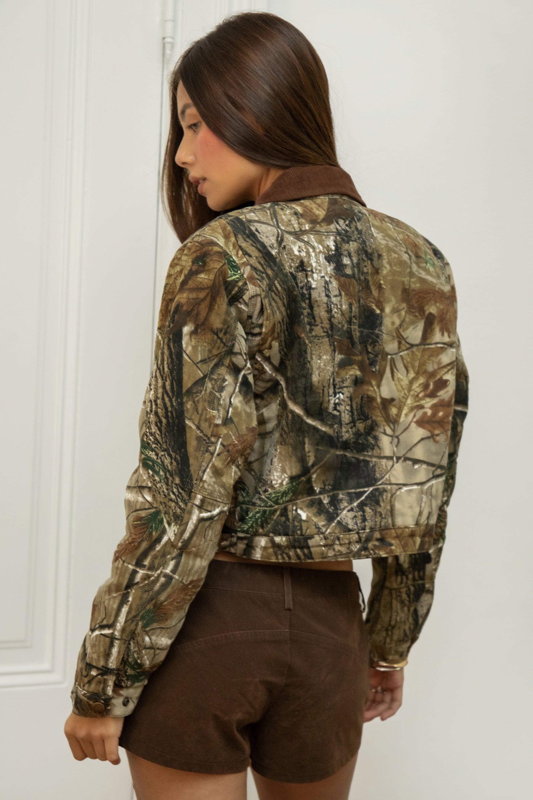 Woman wearing TIC TOC TT32018-OAKLYN OAKLYN camo print cropped jacket featuring an allover camo twill print, front zip closure, contrast brown corduroy collar, two side pockets, a chest zip pocket, and snap button cuffs with side buckle strap detail, styled over a simple top with denim for a western-inspired everyday outdoor look.