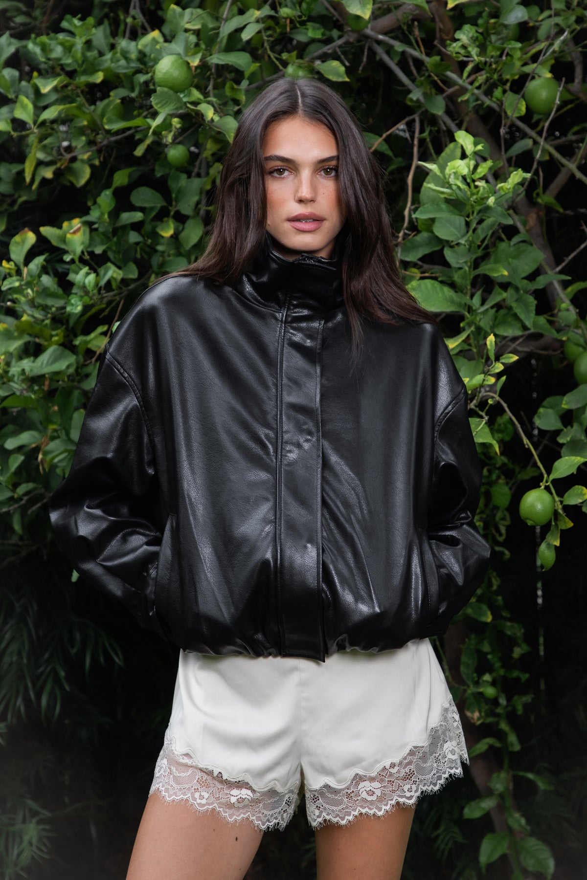 Woman wearing ANAIS oversized PU leather bomber jacket featuring smooth vegan leather fabric with a subtle glossy finish, oversized relaxed fit, cropped bomber silhouette, wide stand collar with snap button detailing, front zipper closure with snap button storm flap, dropped shoulders, adjustable snap button cuffs, elasticated gathered hem, and smooth taffeta lining.