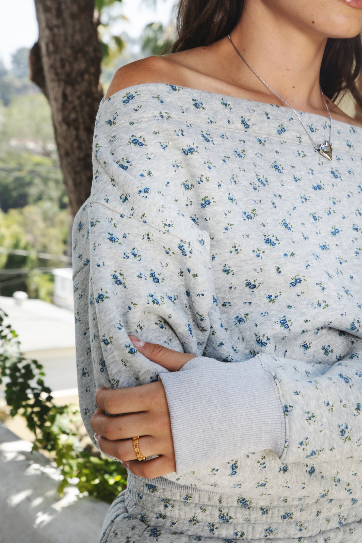 Woman wearing LILIETTE floral off shoulder semi crop sweatshirt featuring soft brushed cotton blend jersey fabric, delicate tiny blue floral print, wide relaxed off shoulder neckline, drop shoulder fit, long sleeves, wide ribbed cuffs, and wide ribbed waistband for a gently cropped matching set look.