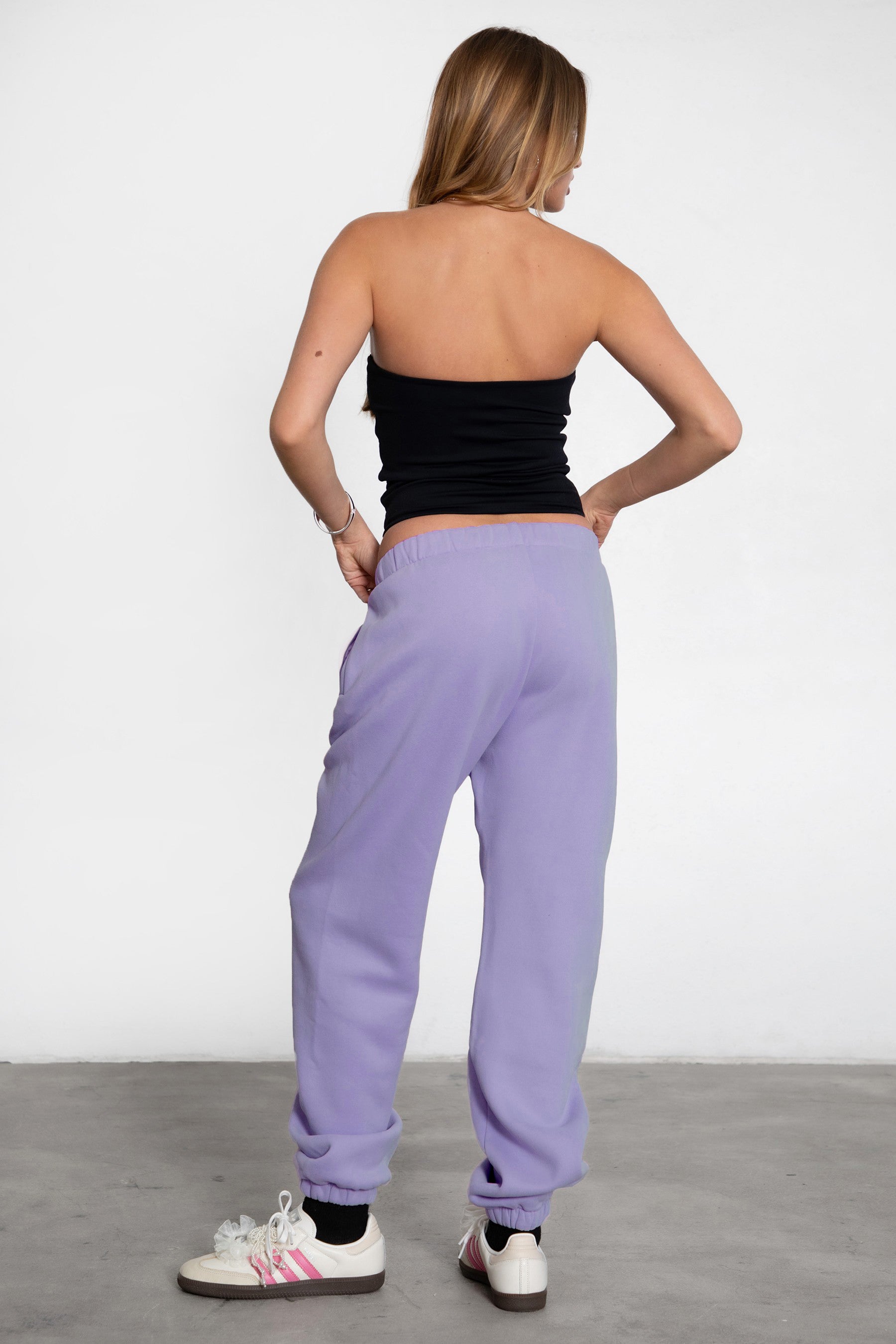 A person wearing high-waisted bright lavender sweatpants with an elastic waistband and side pockets, paired with a black top and white sneakers