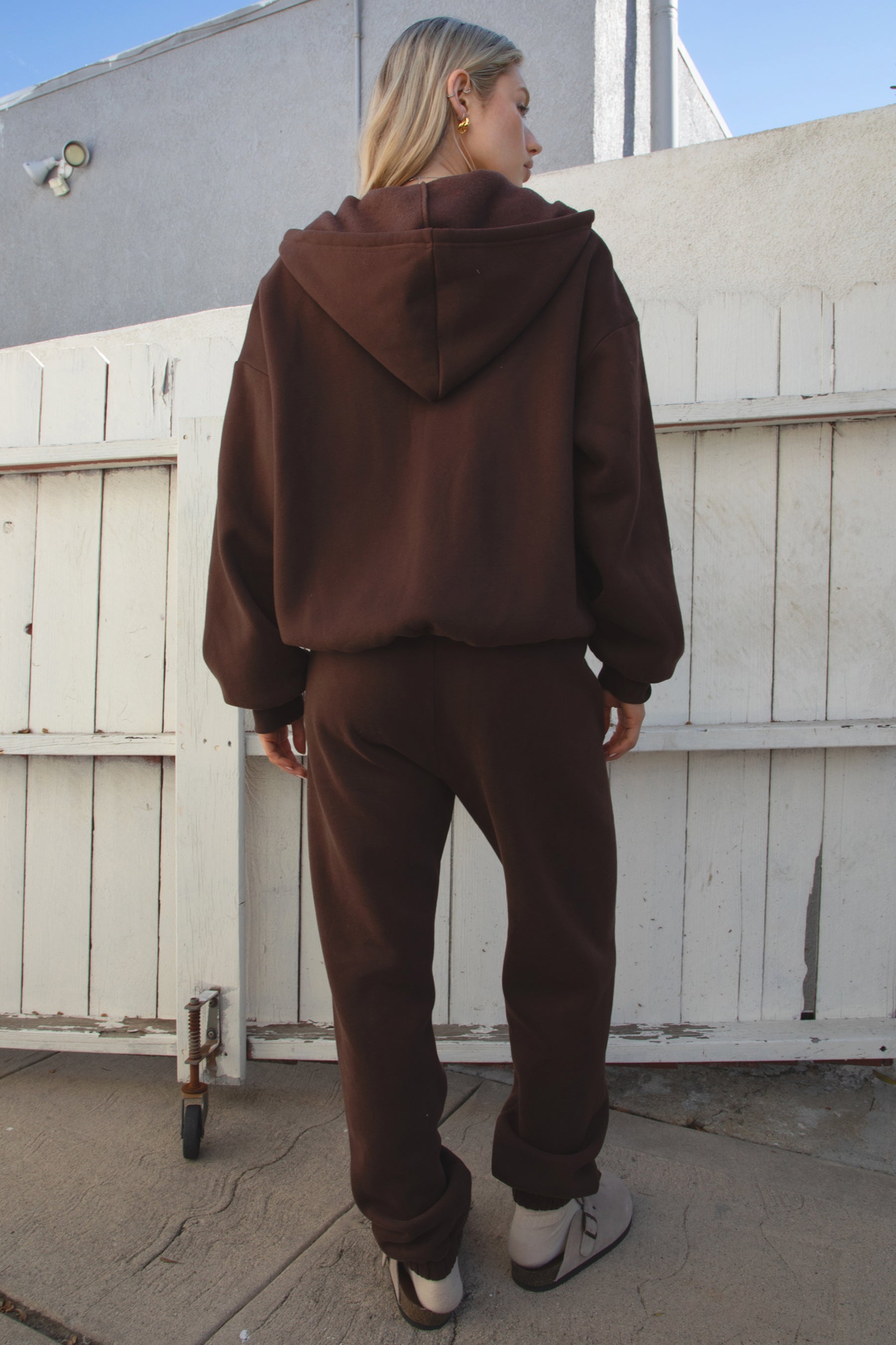 Person wearing a brown tracksuit standing in front of a white wall.