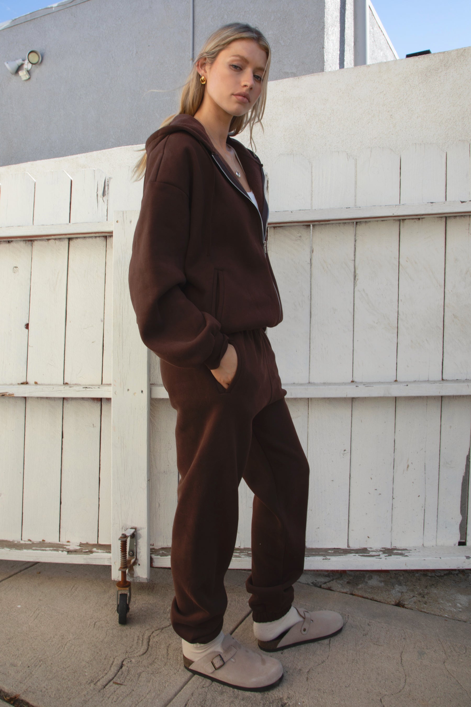 Person wearing a brown tracksuit standing against a white wooden fence.