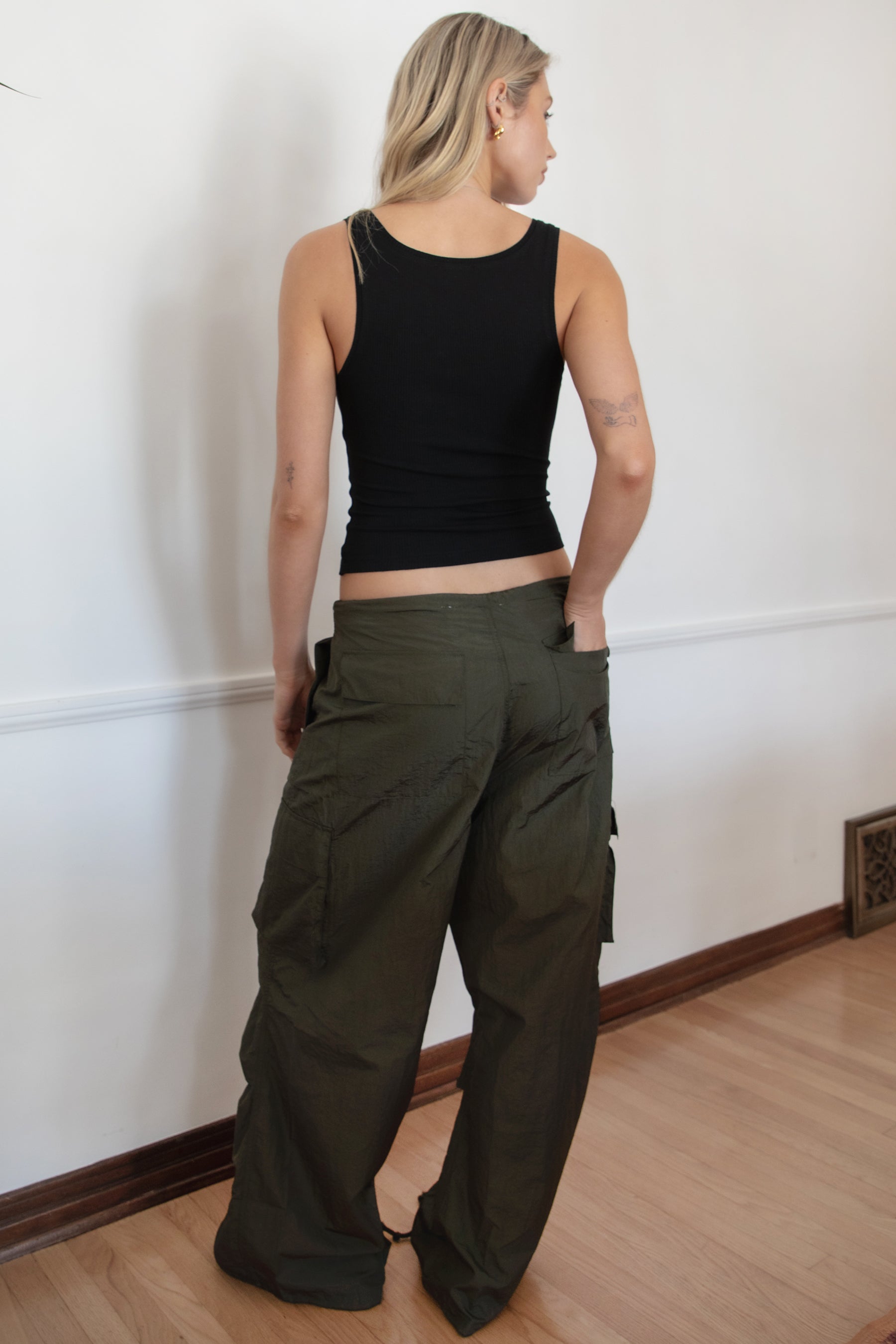Woman wearing a black tank top and TIC TOC PH29131 TOMY olive crinkled nylon parachute cargo pants with adjustable drawstring waistband and wide-leg silhouette against a white wall.