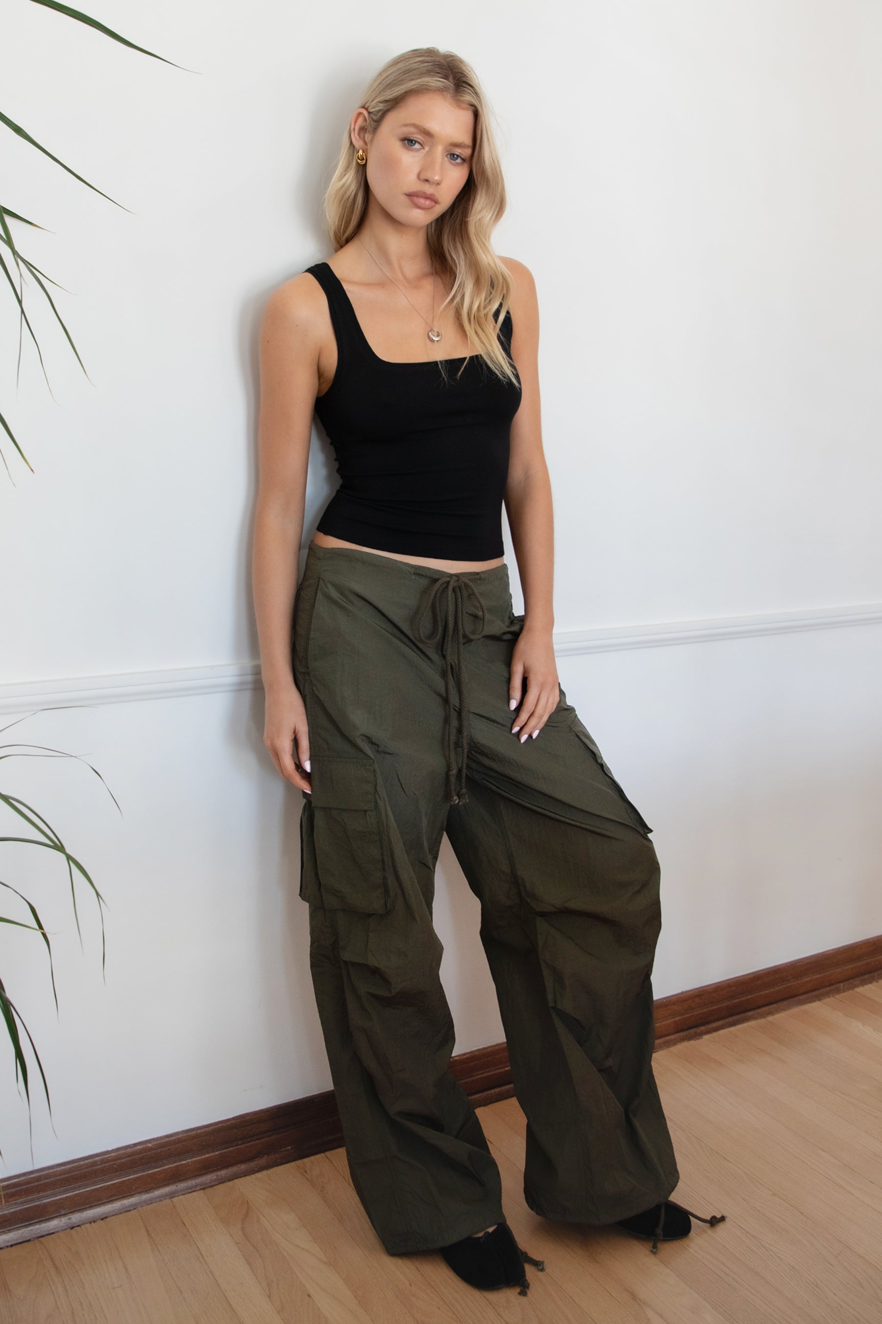 Woman wearing a black tank top and TIC TOC PH29131 TOMY olive crinkled nylon parachute cargo pants with adjustable drawstring waistband and wide-leg silhouette against a white wall.