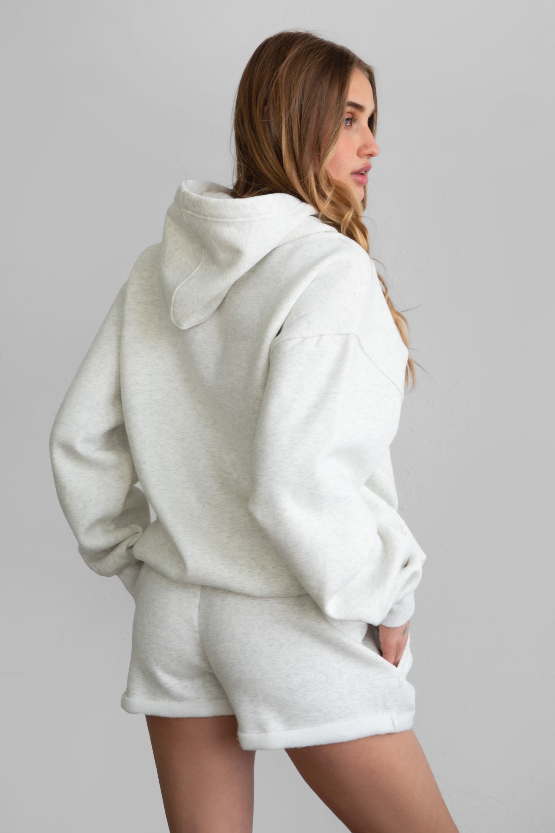 Woman wearing the TIC TOC PJ31004 CARLY heather white cloud fleece low rise shorts featuring an elastic waistband with drawcord, deep side pockets, and raw cut hemline with a matching hoodie