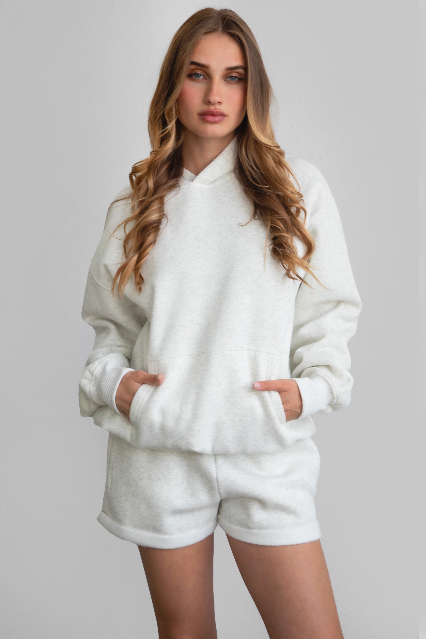 Woman wearing the TIC TOC PJ31004 CARLY heather white cloud fleece low rise shorts featuring an elastic waistband with drawcord, deep side pockets, and raw cut hemline with a matching hoodie