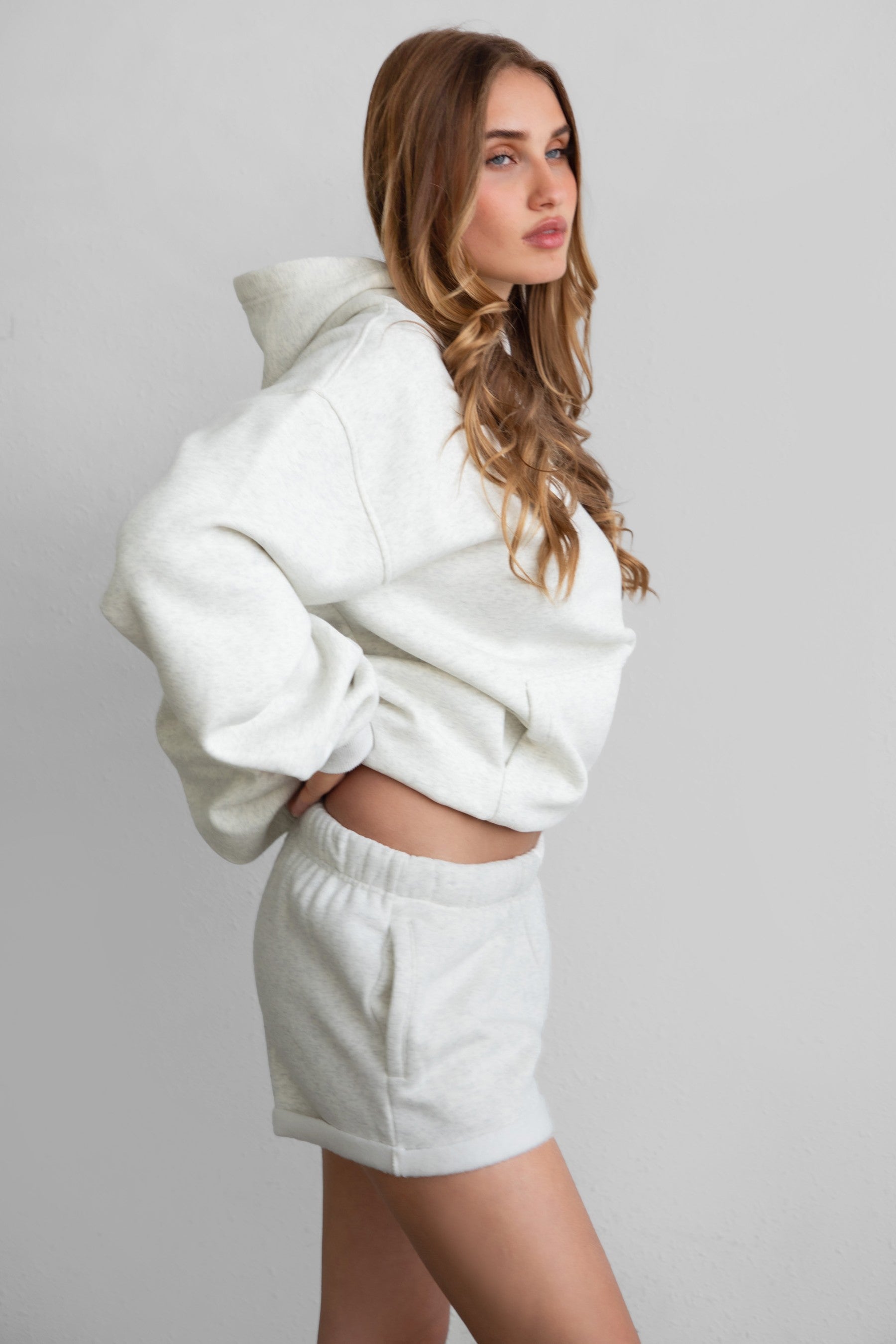Woman wearing the TIC TOC PJ31004 CARLY heather white cloud fleece low rise shorts featuring an elastic waistband with drawcord, deep side pockets, and raw cut hemline with a matching hoodie