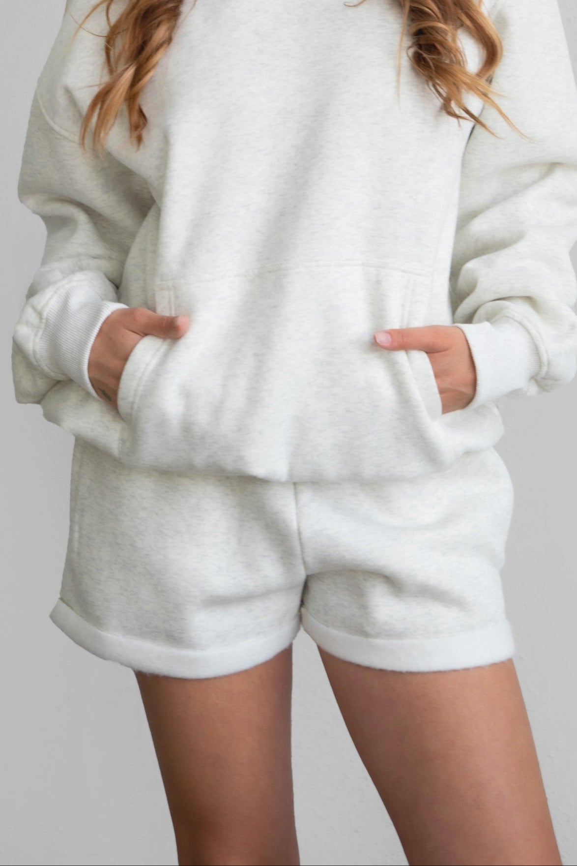 Woman wearing the TIC TOC PJ31004 CARLY heather white cloud fleece low rise shorts featuring an elastic waistband with drawcord, deep side pockets, and raw cut hemline with a matching hoodie