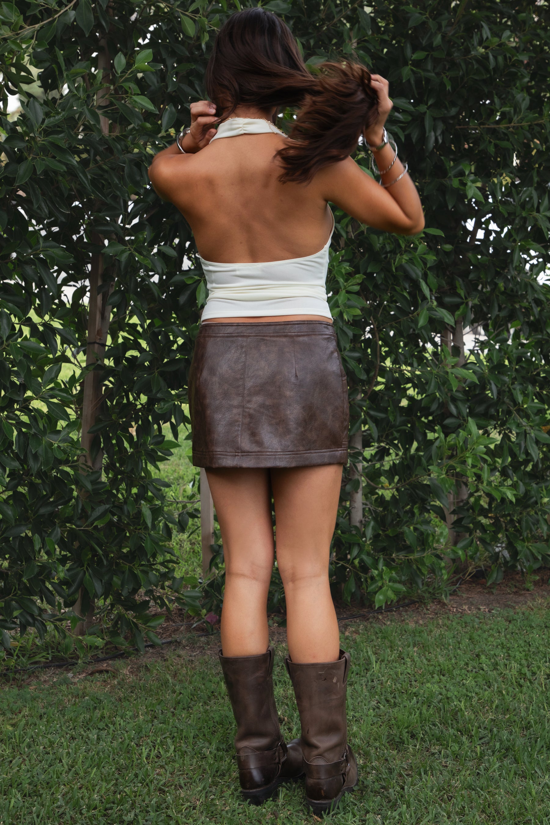 Woman wearing the TIC TOC SH31455 DAYNA brown PU leather low rise mini skirt featuring faux side zippers, button closure, and functional pockets in stretch leather fabric paired with a cream cowl neck top and knee high boots. 